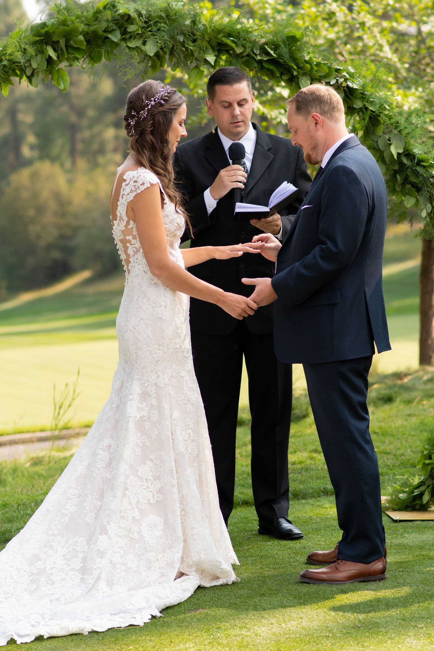 Outdoor wedding ceremony showing ring exchange between couple on grassy lawn with officiant under leafy trees.