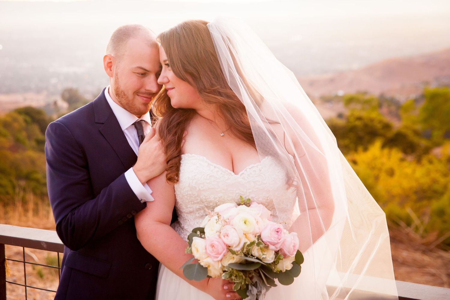 A romantic couple in wedding attire shares an intimate moment during sunset in a scenic outdoor setting.