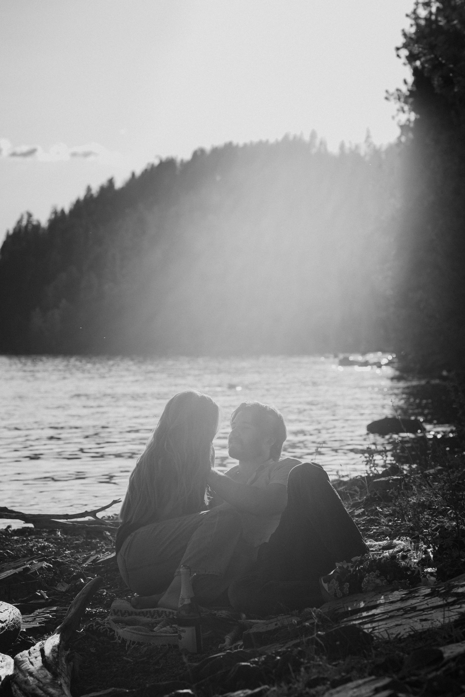 Silhouetted figures sit on a rocky beach at sunset with a misty forest coastline in the background.