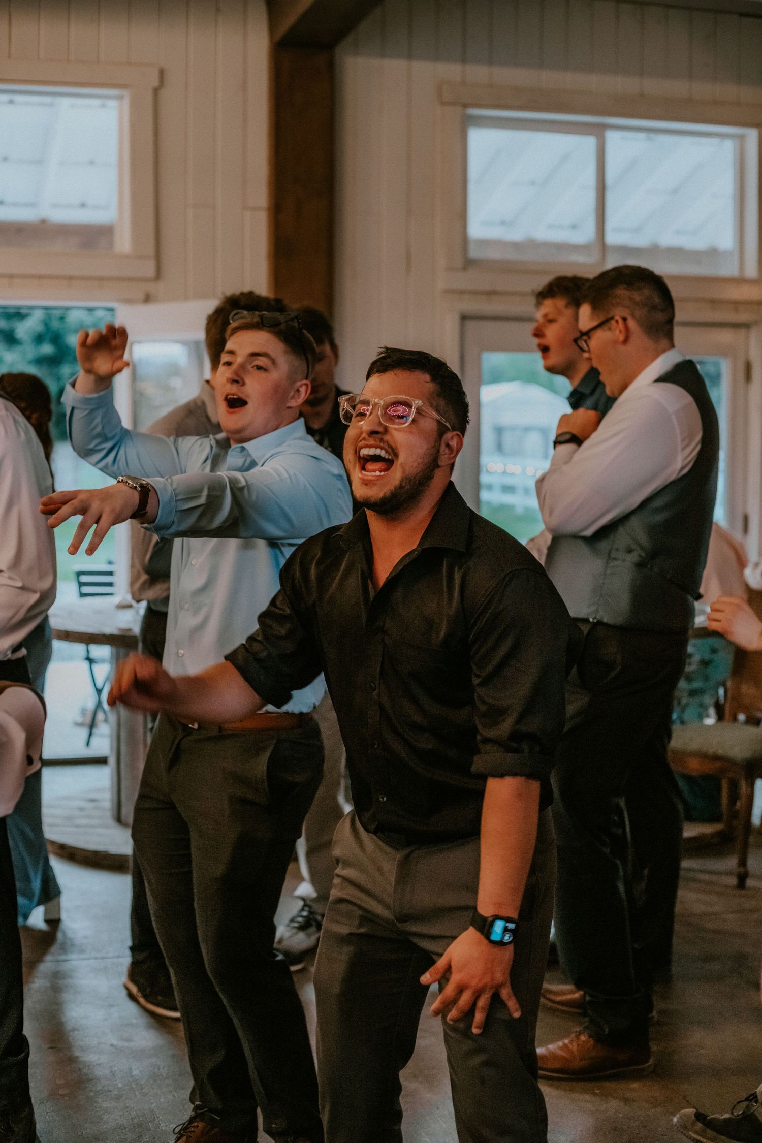 Wedding groomsmen laugh and drink together during celebratory wedding reception toast at indoor venue.