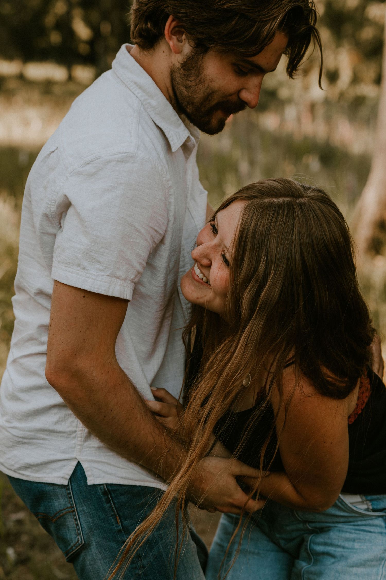 A romantic couple embraces outdoors during golden hour, sharing an intimate moment in casual attire.