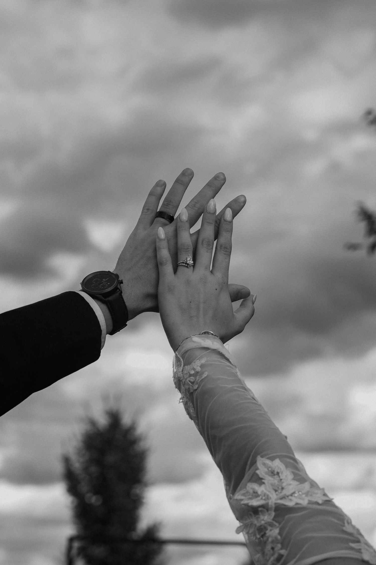 Two hands reach towards the cloudy sky showing wedding rings in dramatic black and white photography.