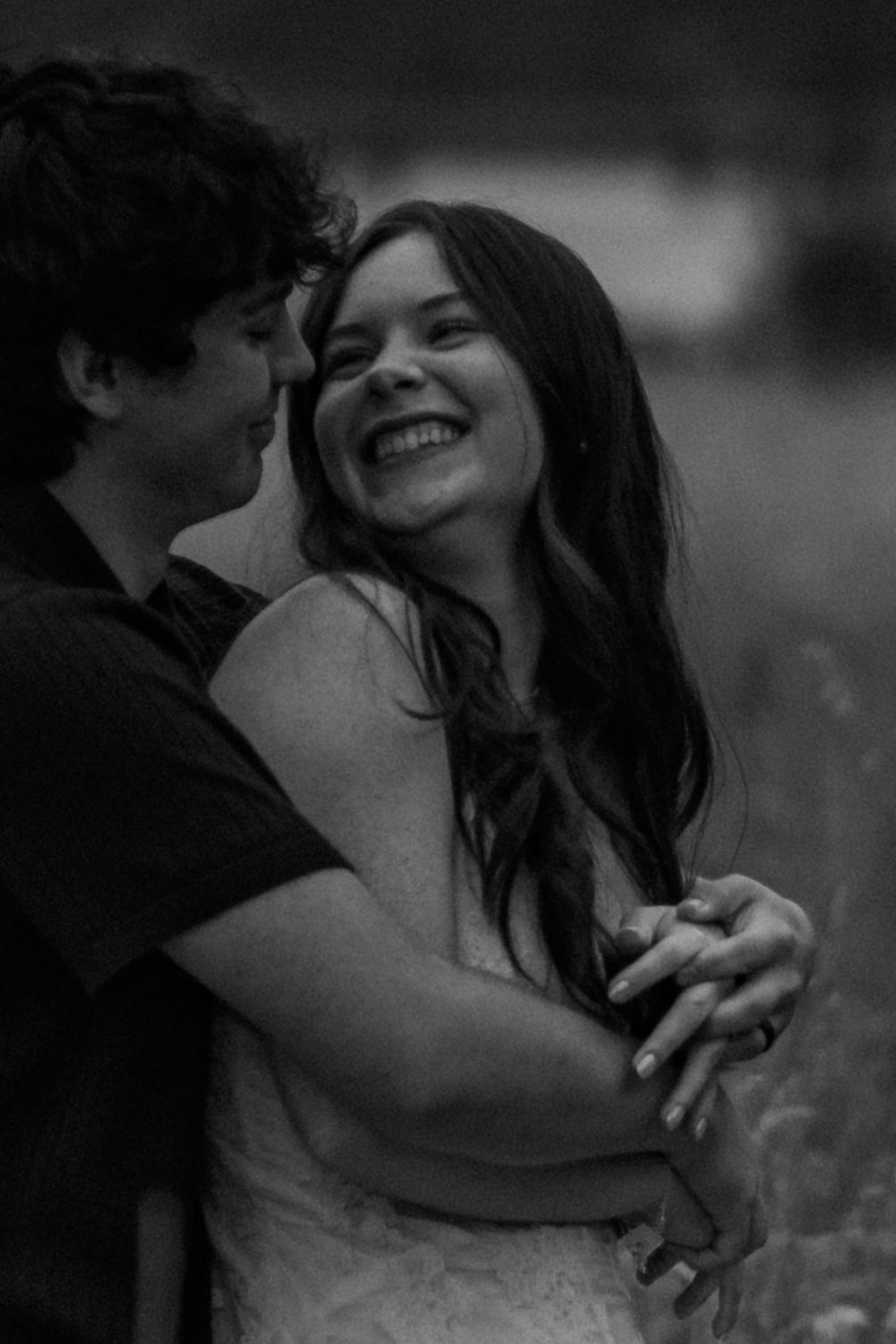 Black and white photo of a happy couple embracing outdoors, woman smiling joyfully as man whispers in her ear.