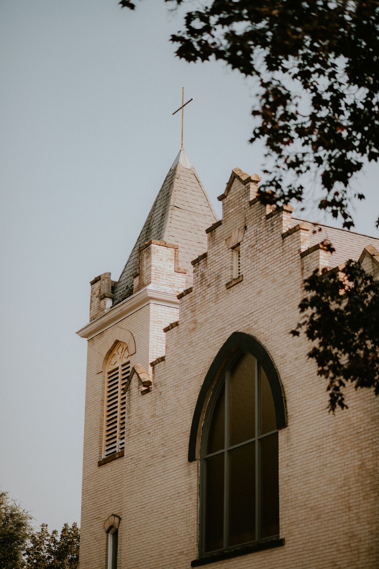 Three gothic church steeples with crosses and arched windows rise against a cloudy sky, framed by tree branches.
