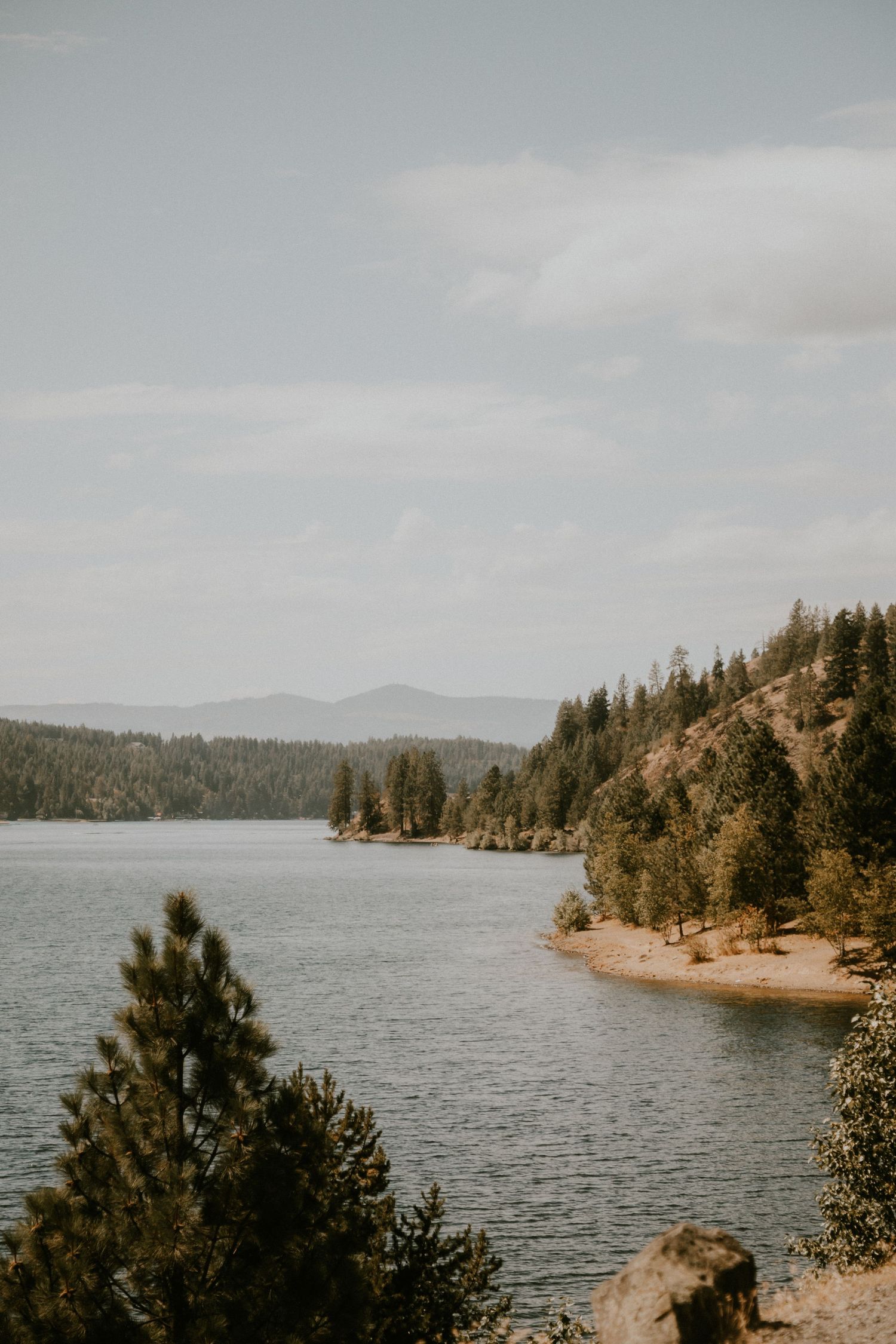 Scenic panoramic view of a lake surrounded by evergreen trees and rocky outcrops under an overcast sky.