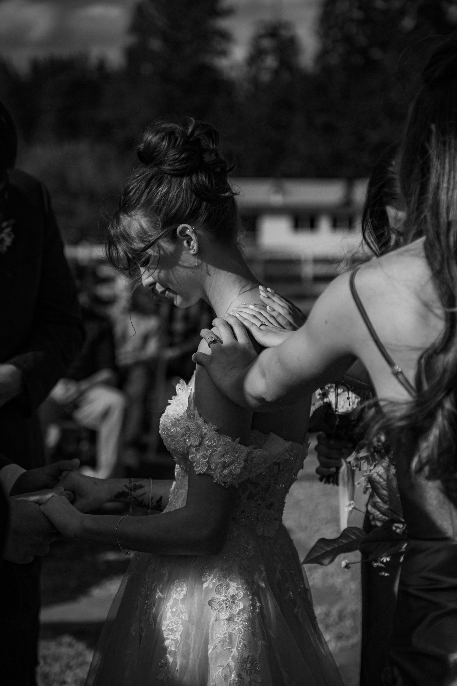 Black and white candid wedding photo showing a bride in a lace dress being helped with final preparations outdoors.