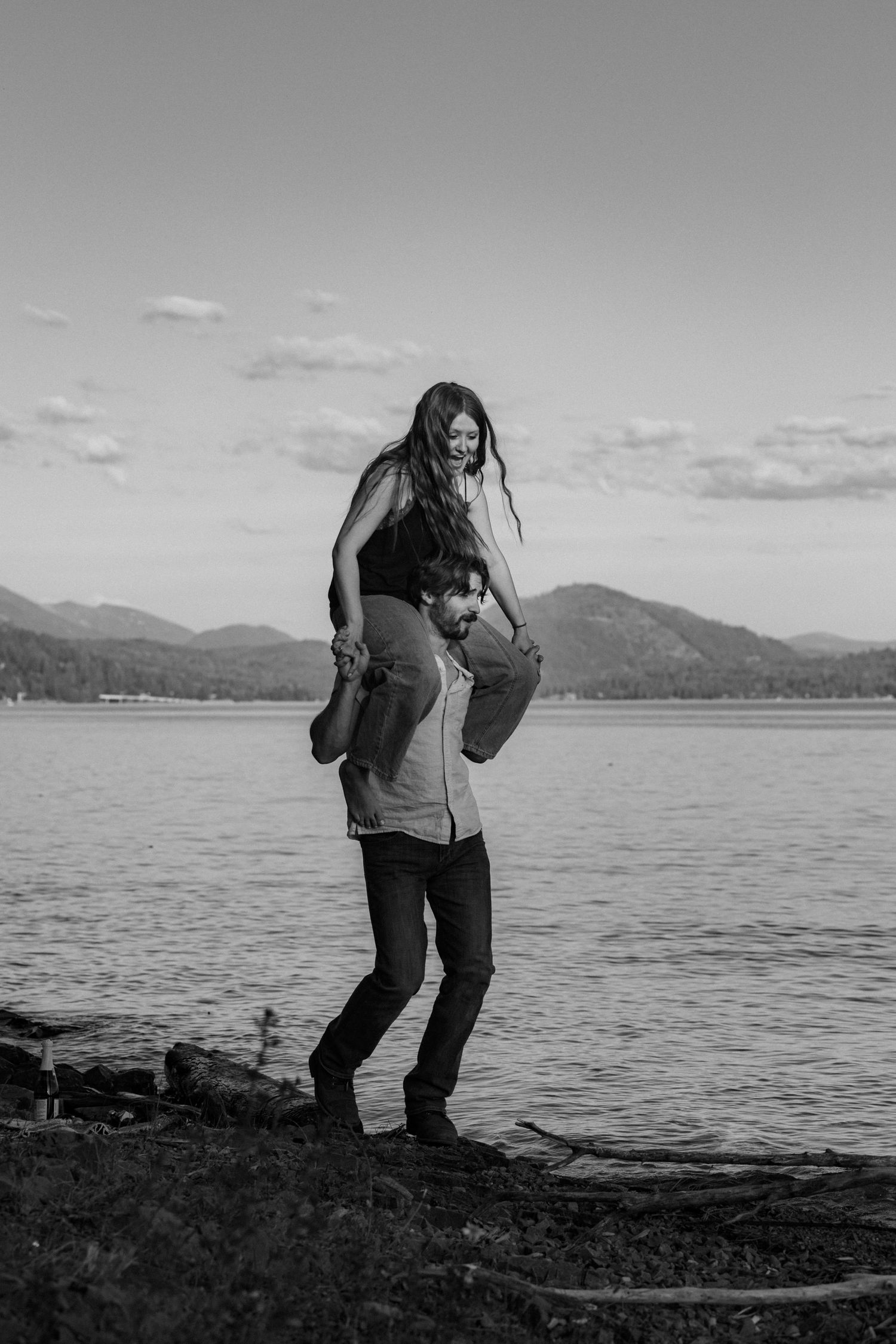 A couple shares a joyful moment by a lake with mountains in the background, captured in black and white photography.