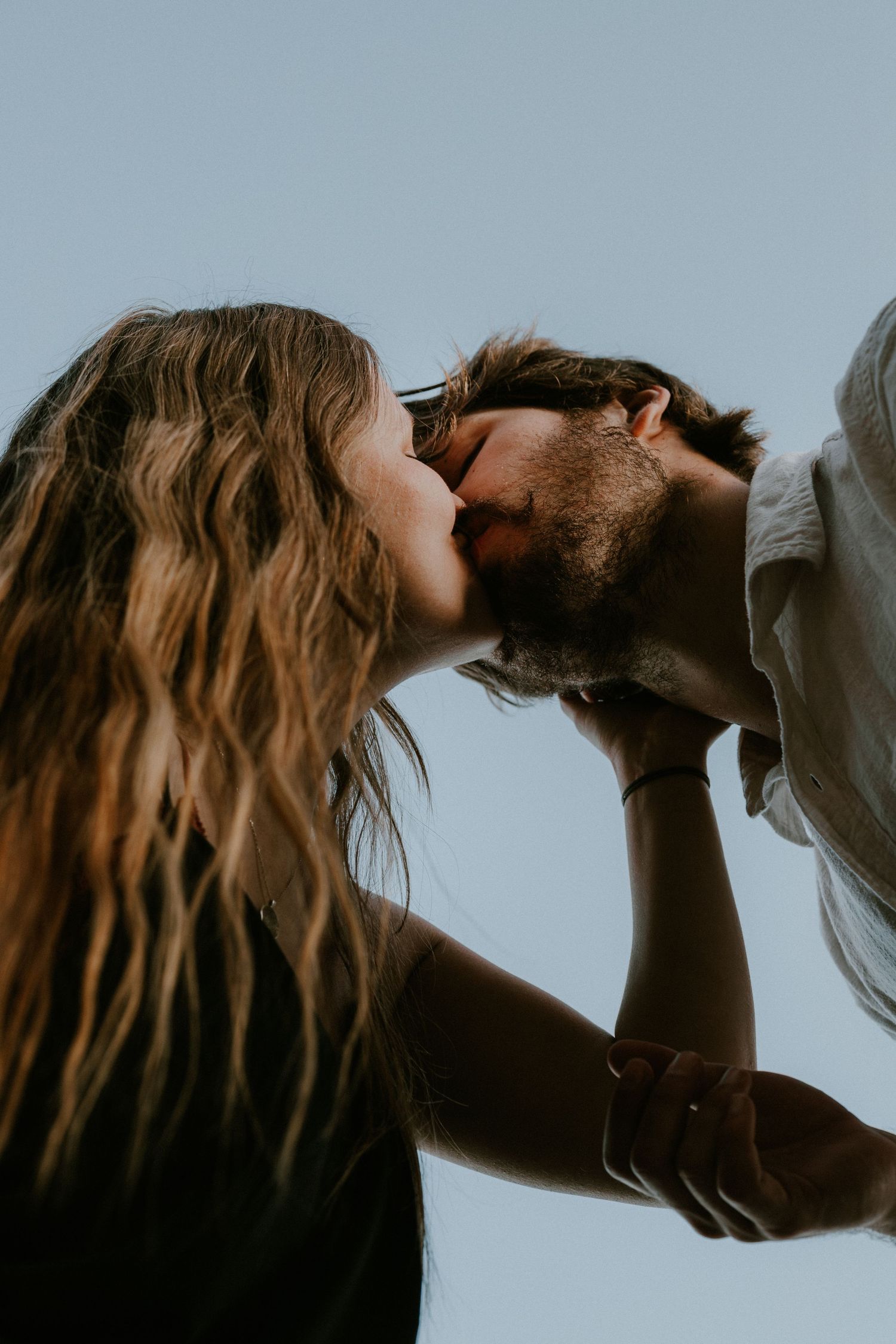 A romantic couple sharing an intimate kiss against a soft blue sky background.