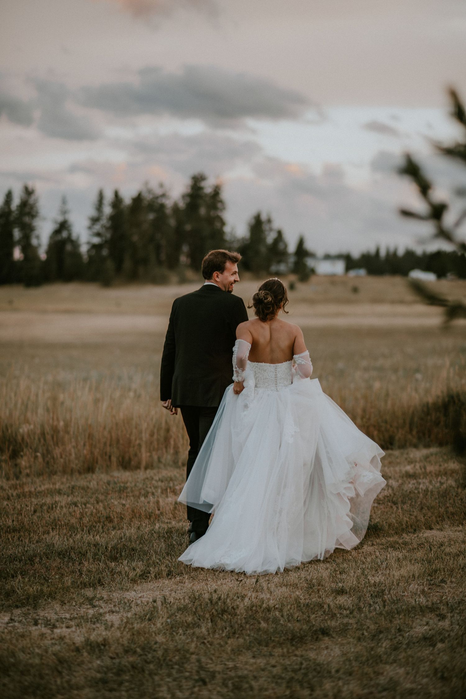 A bride and groom walk together through a serene field at sunset with evergreen trees in the background.