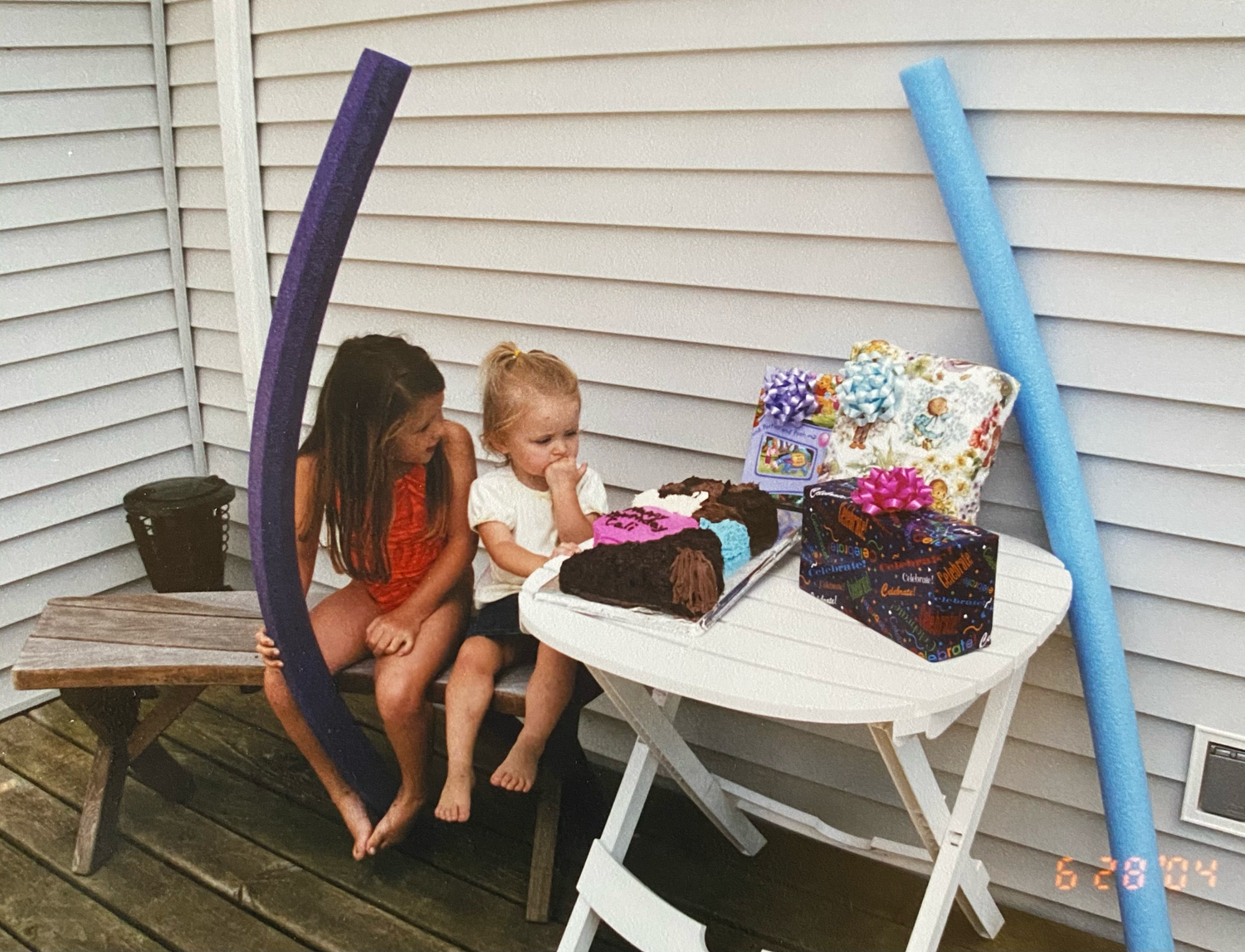 Two people sit on a wooden deck near a white table with pool noodles and swimsuits in the summertime.
