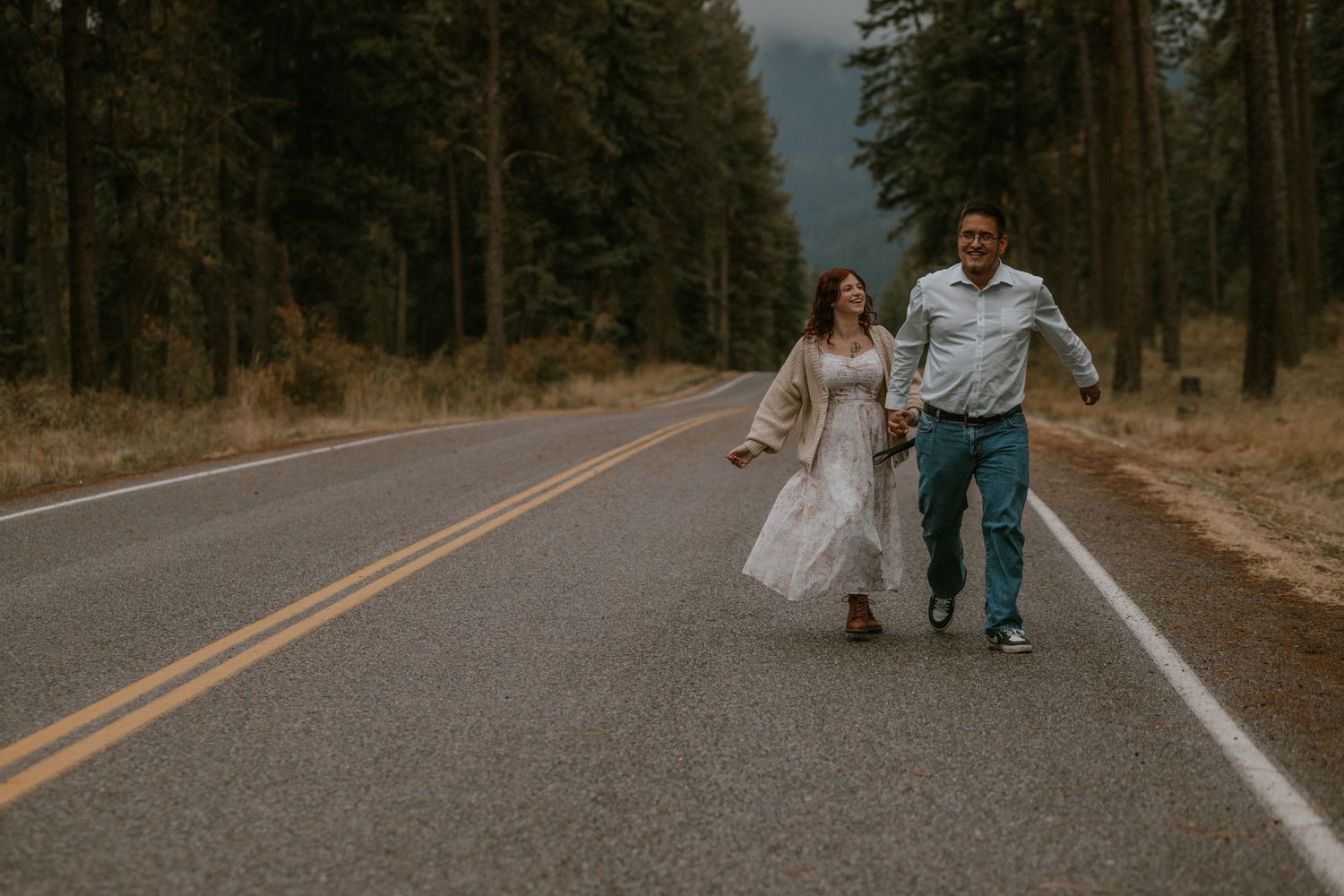 A couple walks hand in hand down a winding mountain road surrounded by tall pine trees at sunset.