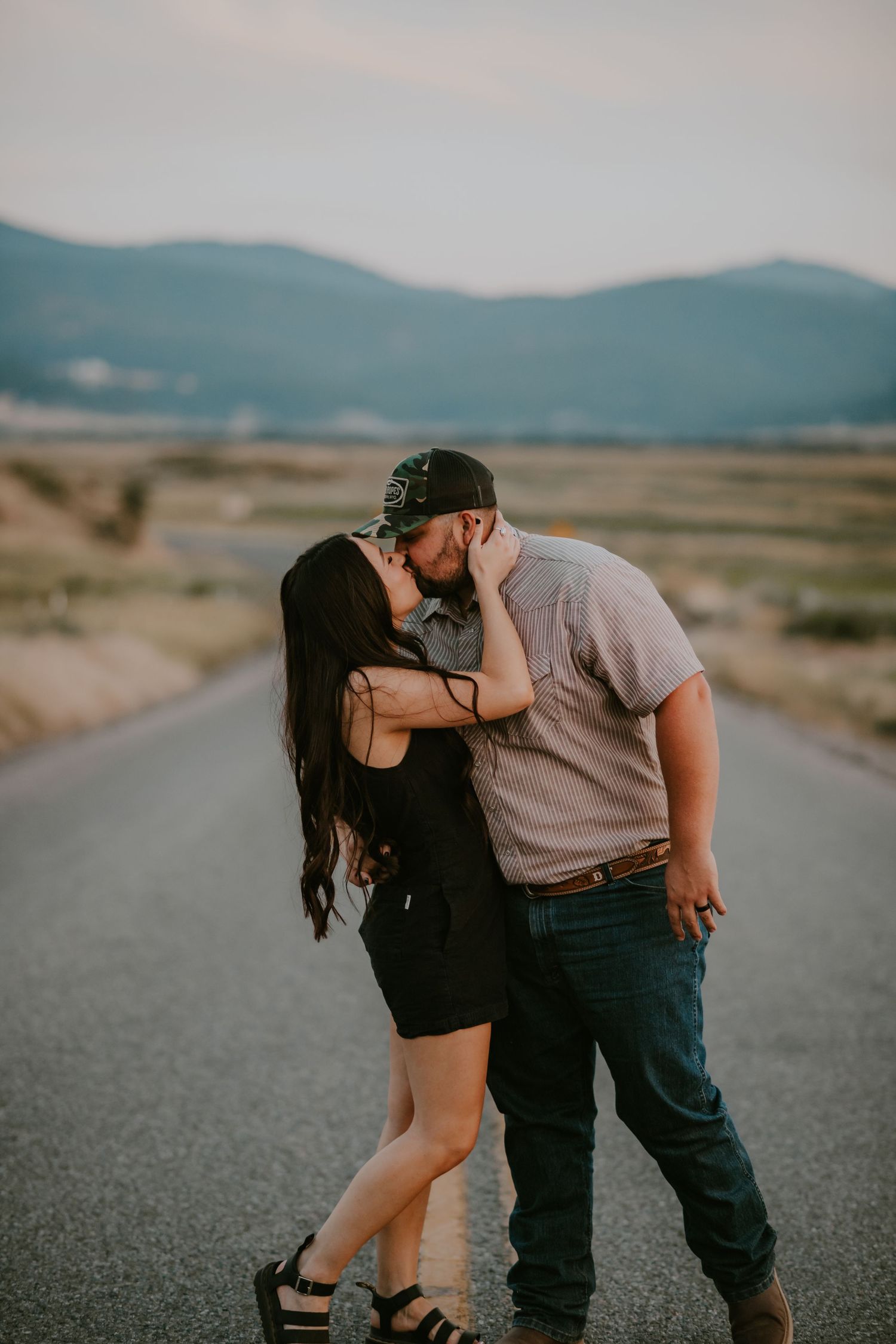 A couple shares a romantic moment on an empty rural road with mountains in the distance.