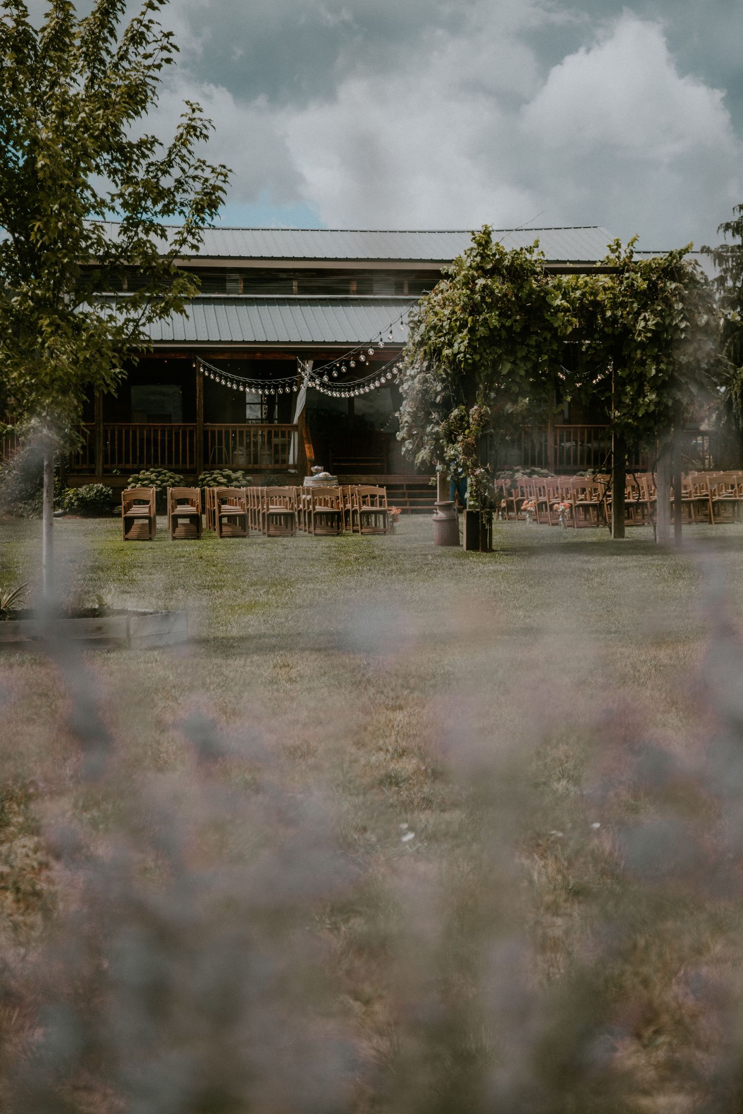 Rustic outdoor wedding ceremony setup with wooden chairs, string lights, and lush greenery beside a barn venue.