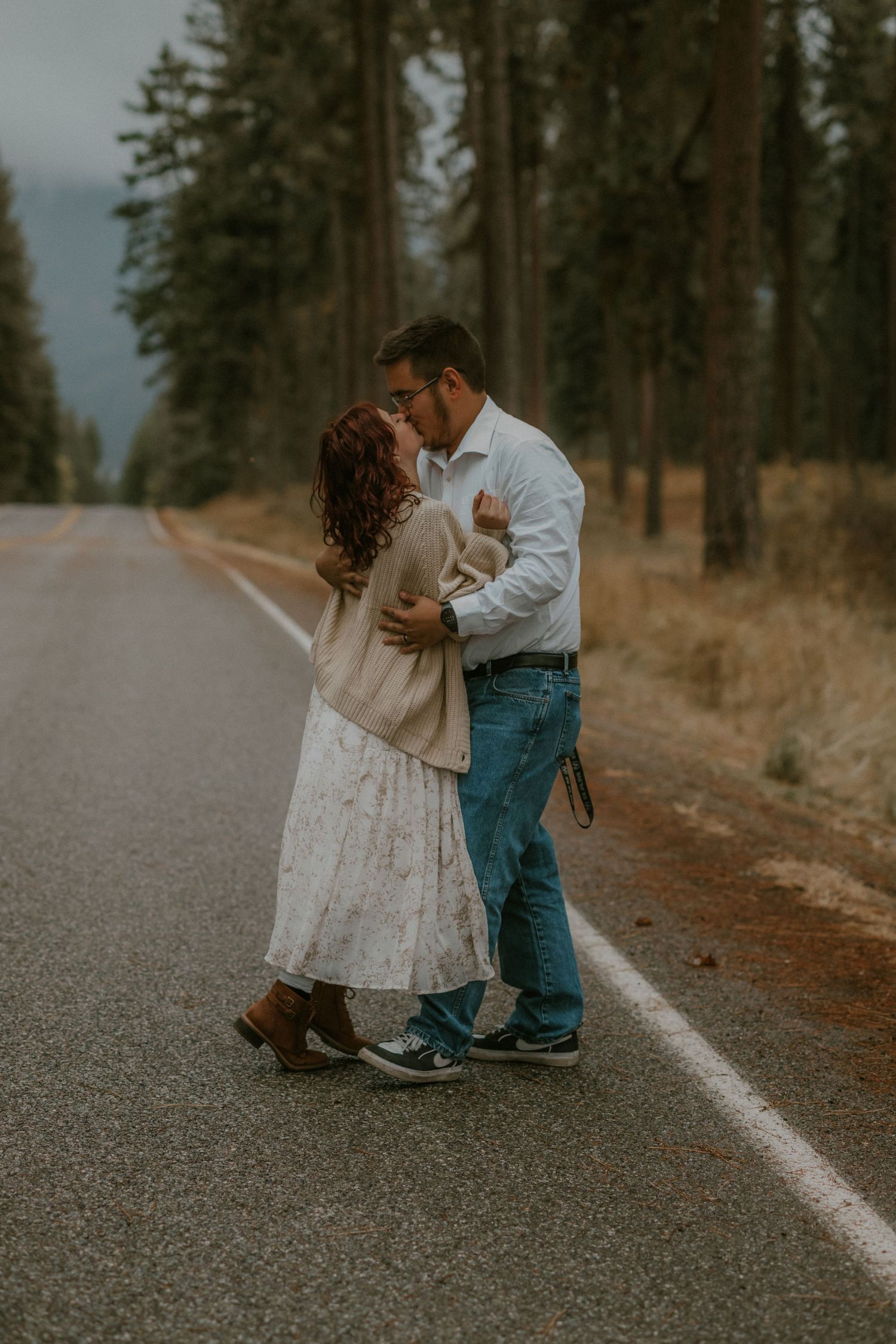 A romantic couple embraces on a forest road during a moody autumn day, surrounded by tall pine trees.
