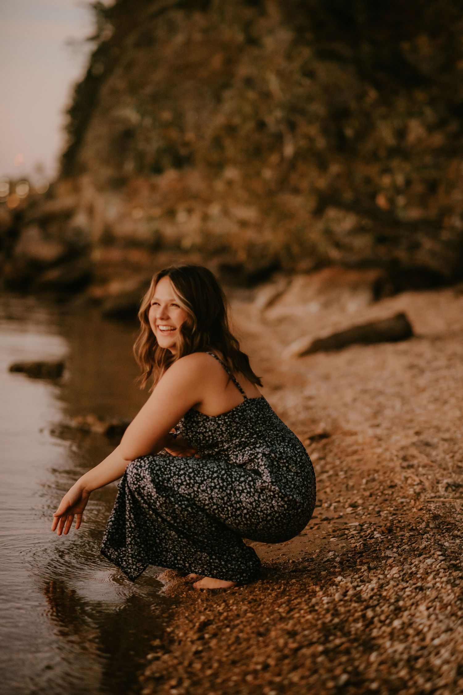 Woman in dark patterned dress posing playfully by rocky shoreline in warm sunset lighting.