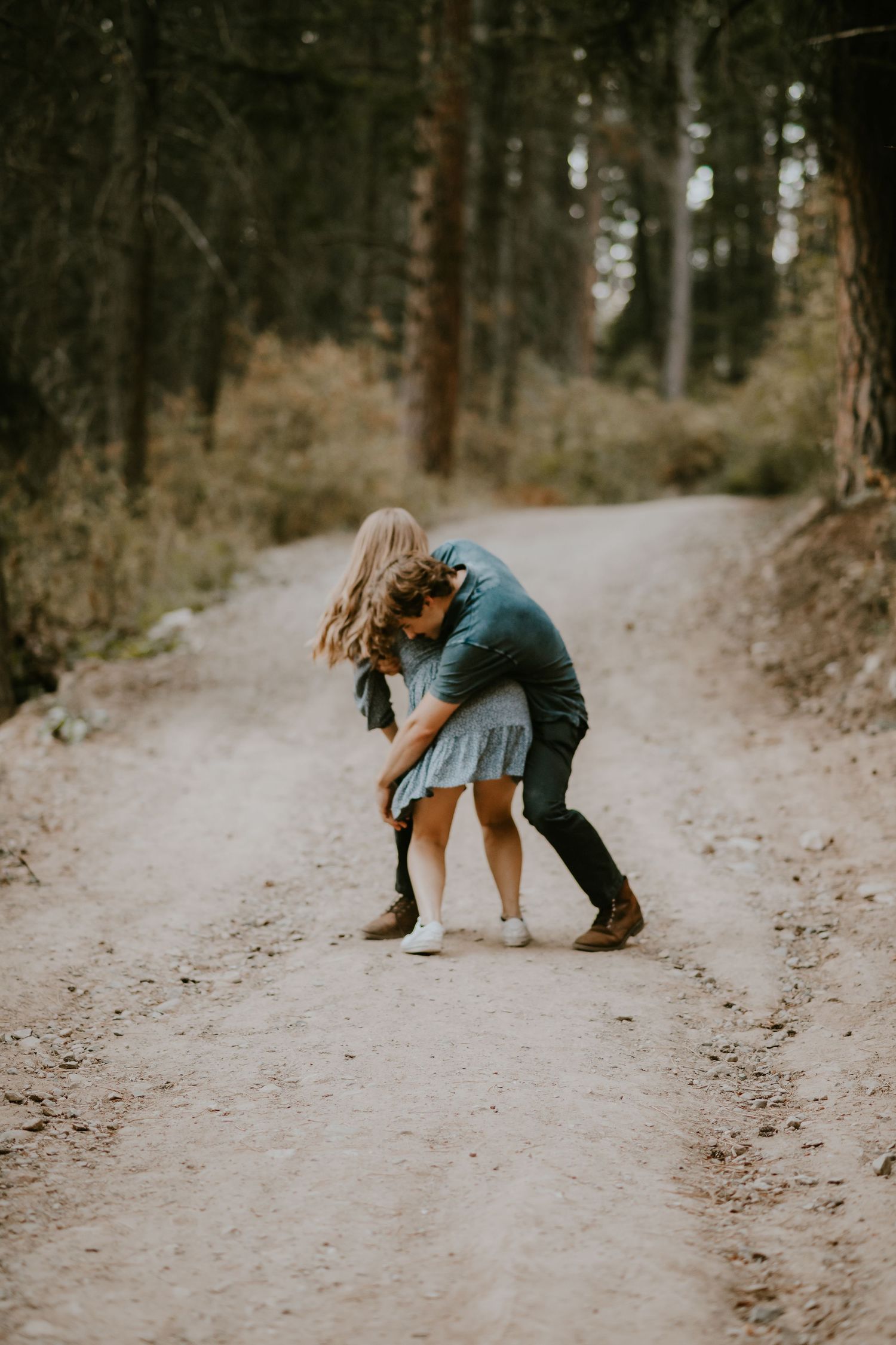 A couple embraces on a dirt path through a wooded forest setting during what appears to be a romantic photoshoot.