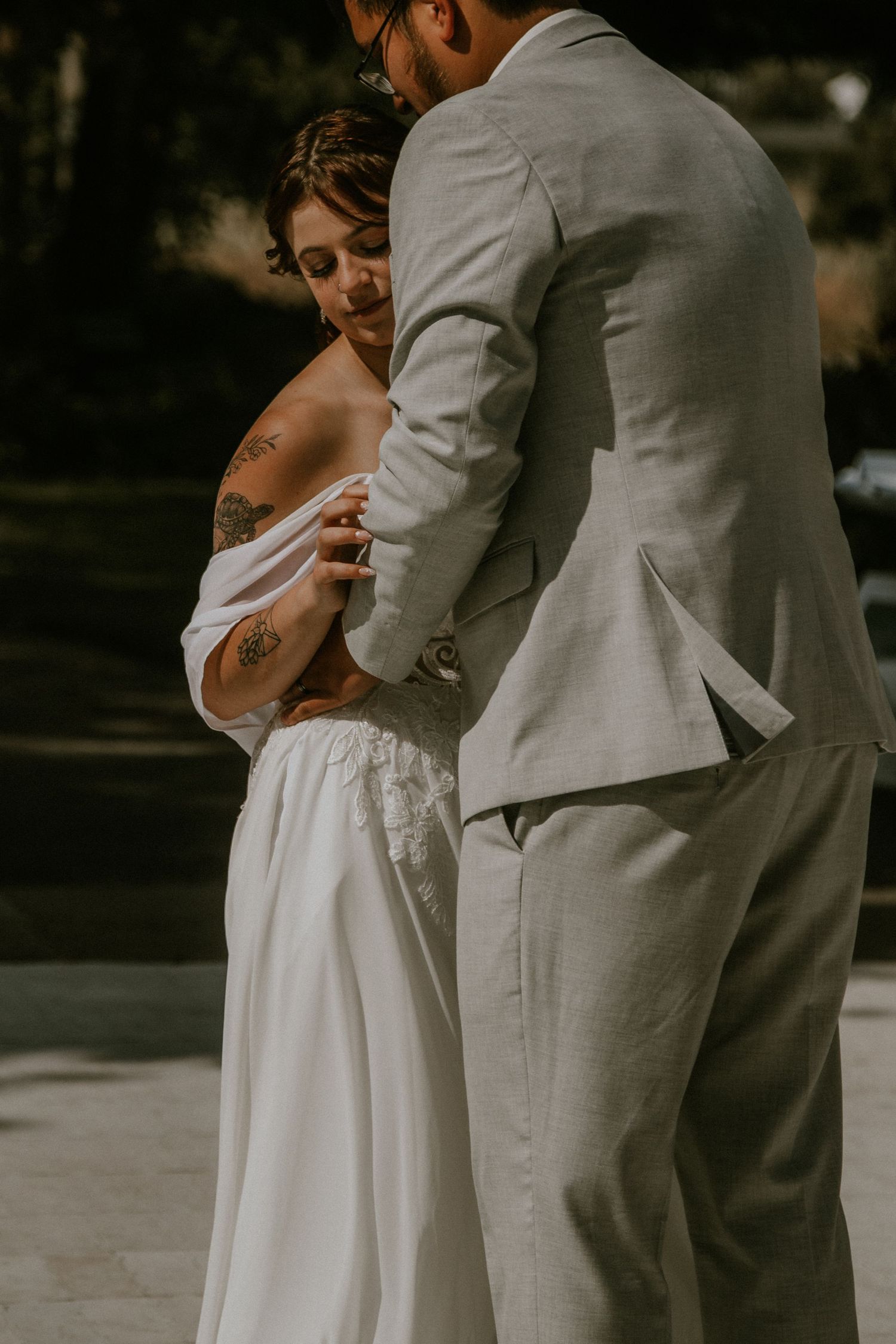 A bride in a white off-shoulder gown with tattoos embraces a groom in a light gray linen suit outdoors.