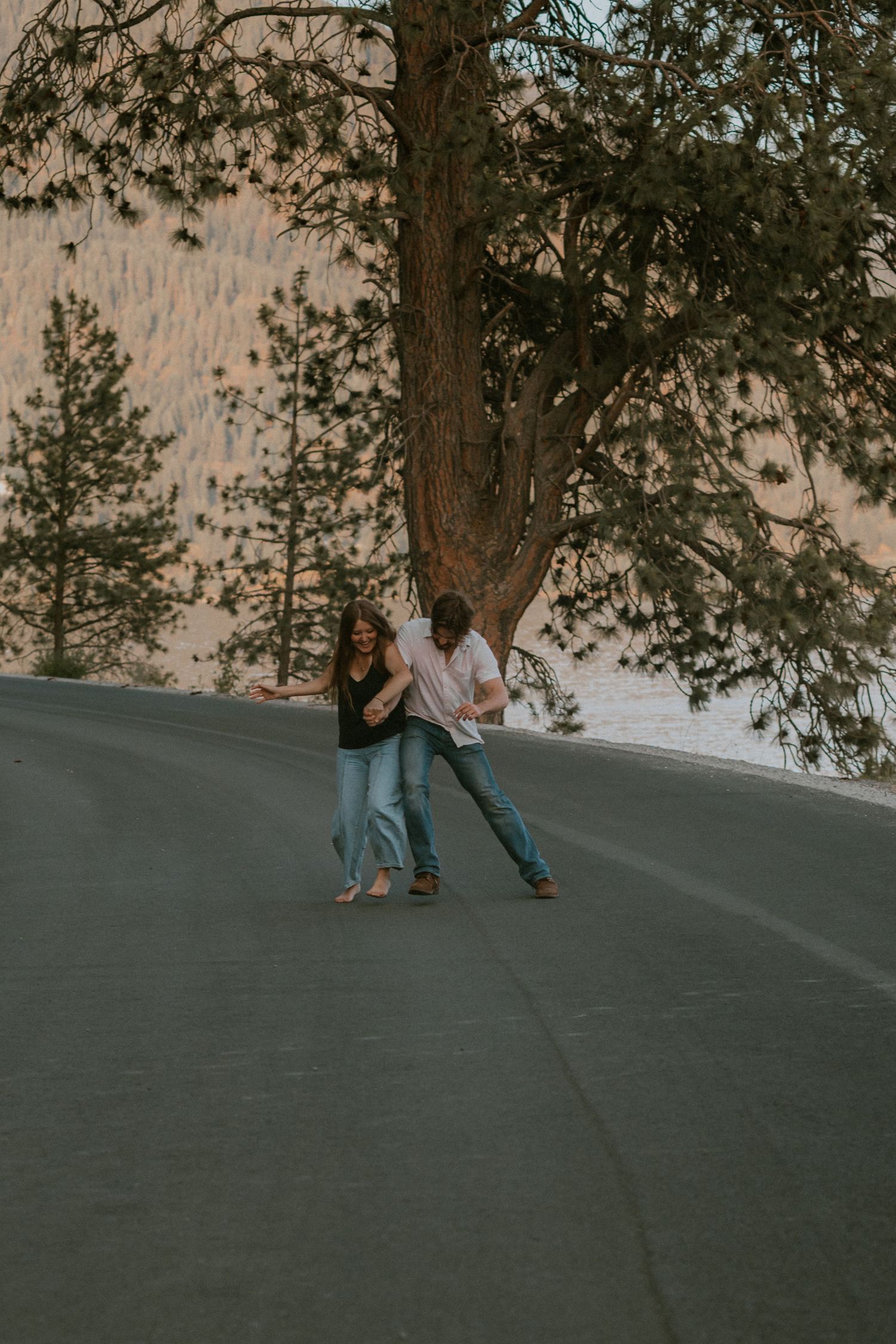 A couple shares a romantic moment on a curved road surrounded by trees at dusk.