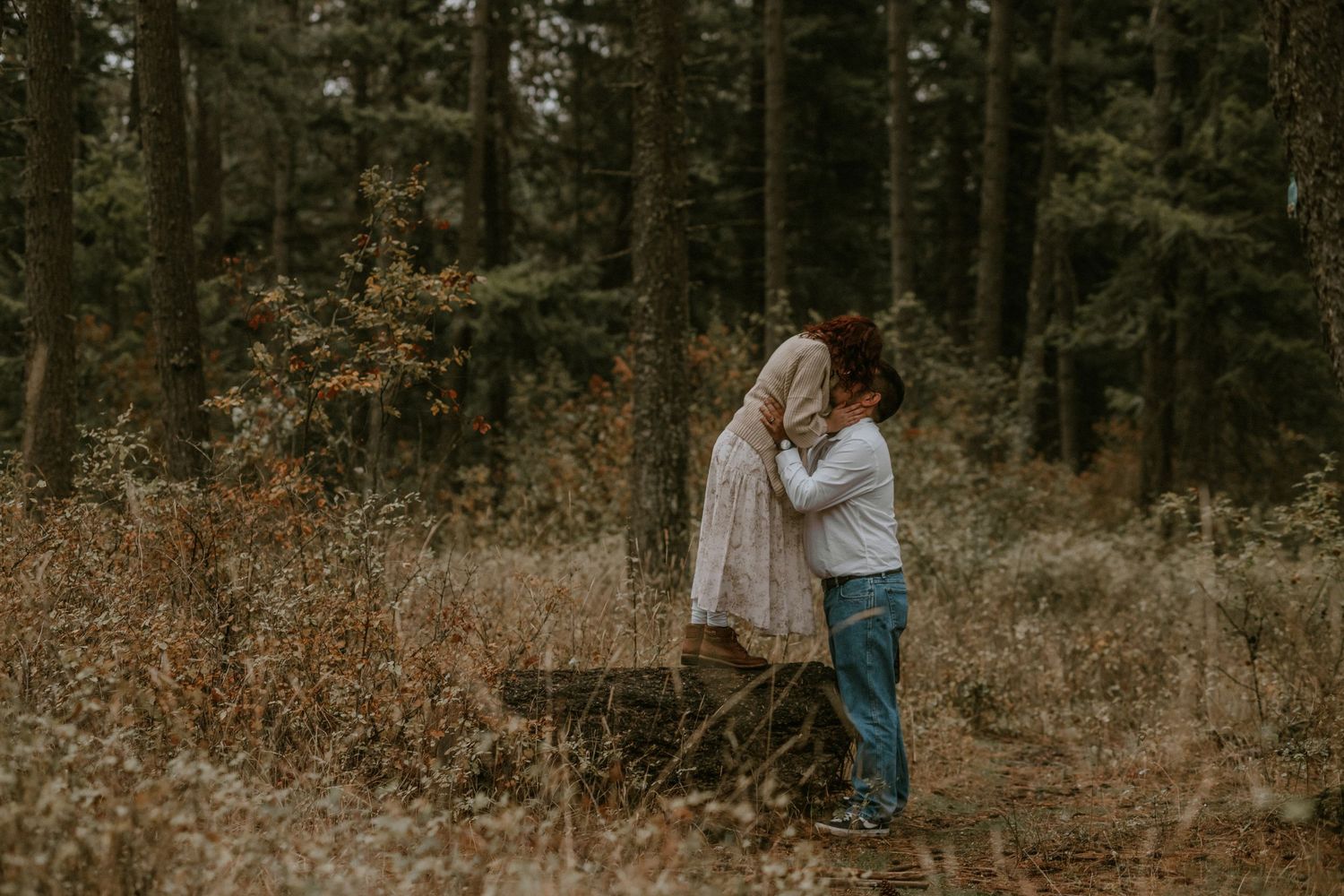 A couple embraces in a romantic forest setting with autumn foliage and soft natural lighting.