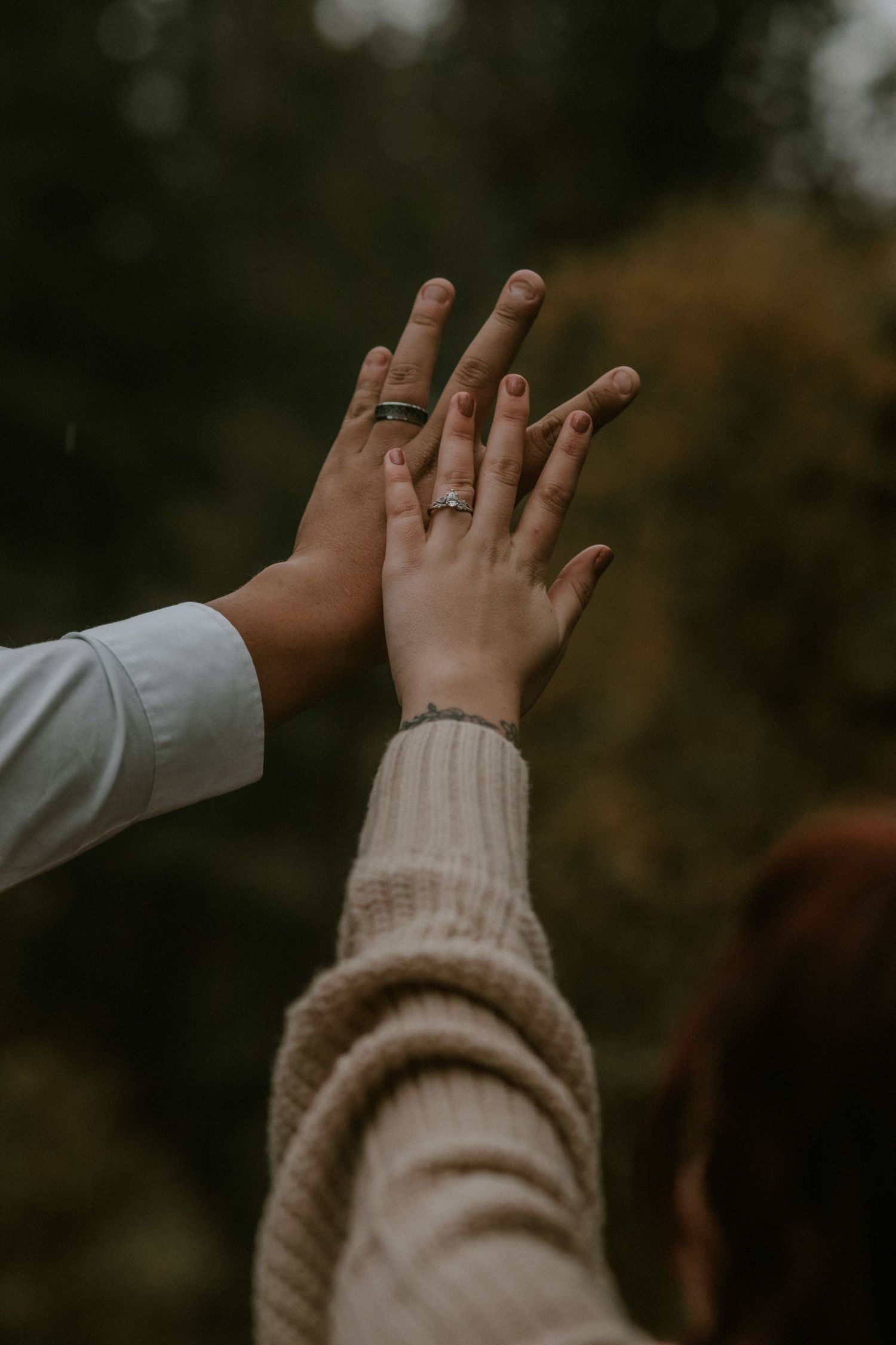 Two hands wearing engagement rings touch together against a blurred outdoor autumn background.