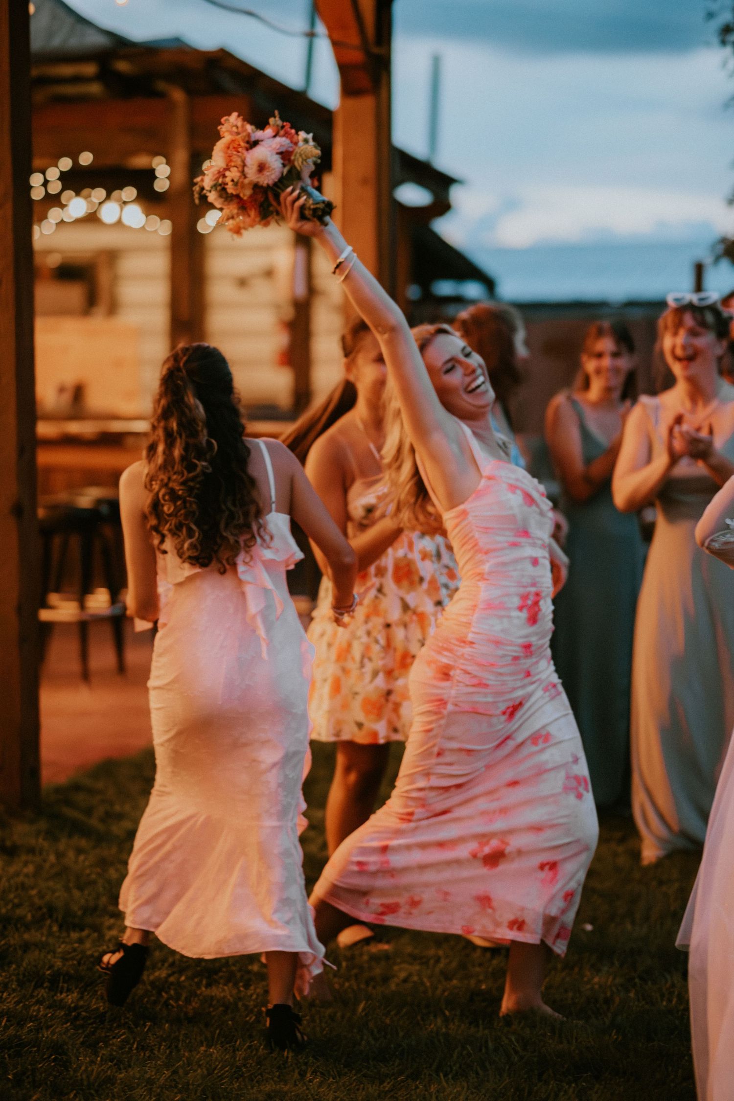 Wedding party guests dancing joyfully at an outdoor evening reception with warm lighting and a floral bouquet toss.