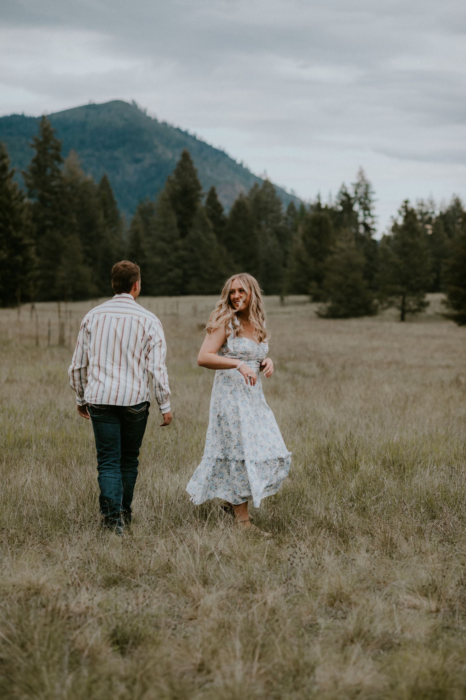 A couple walks together in a meadow with mountains and evergreen forest in the background on an overcast day.