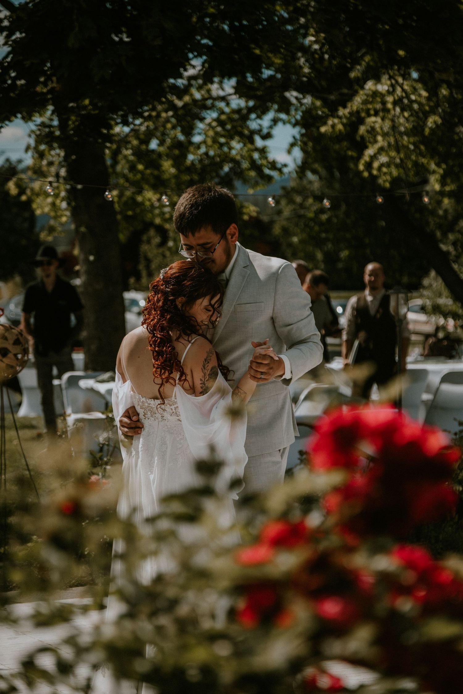 Bride and groom share a romantic embrace outdoors at a sunlit garden wedding ceremony with red roses in the foreground.