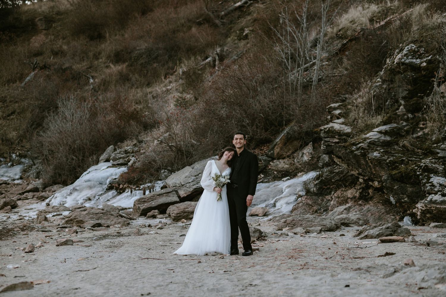 A couple in formal wedding attire walks along a rocky winter landscape through fresh snow.