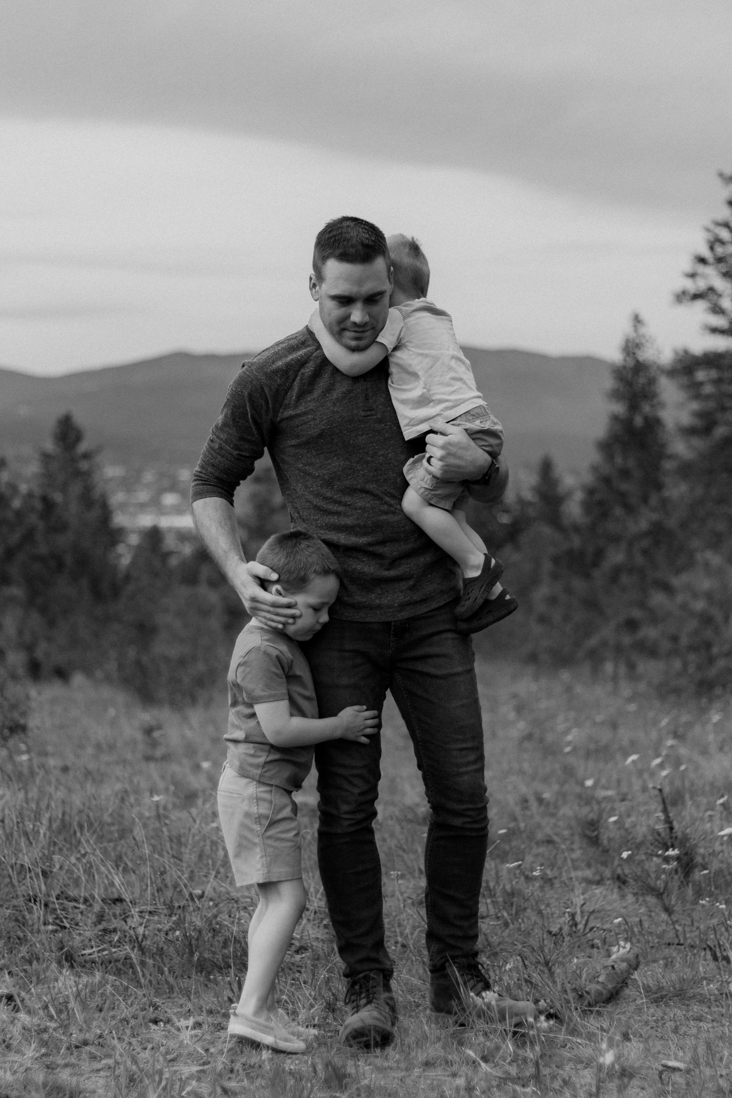 A parent and child share a tender moment in a mountain meadow with evergreen trees and rolling hills in the background.