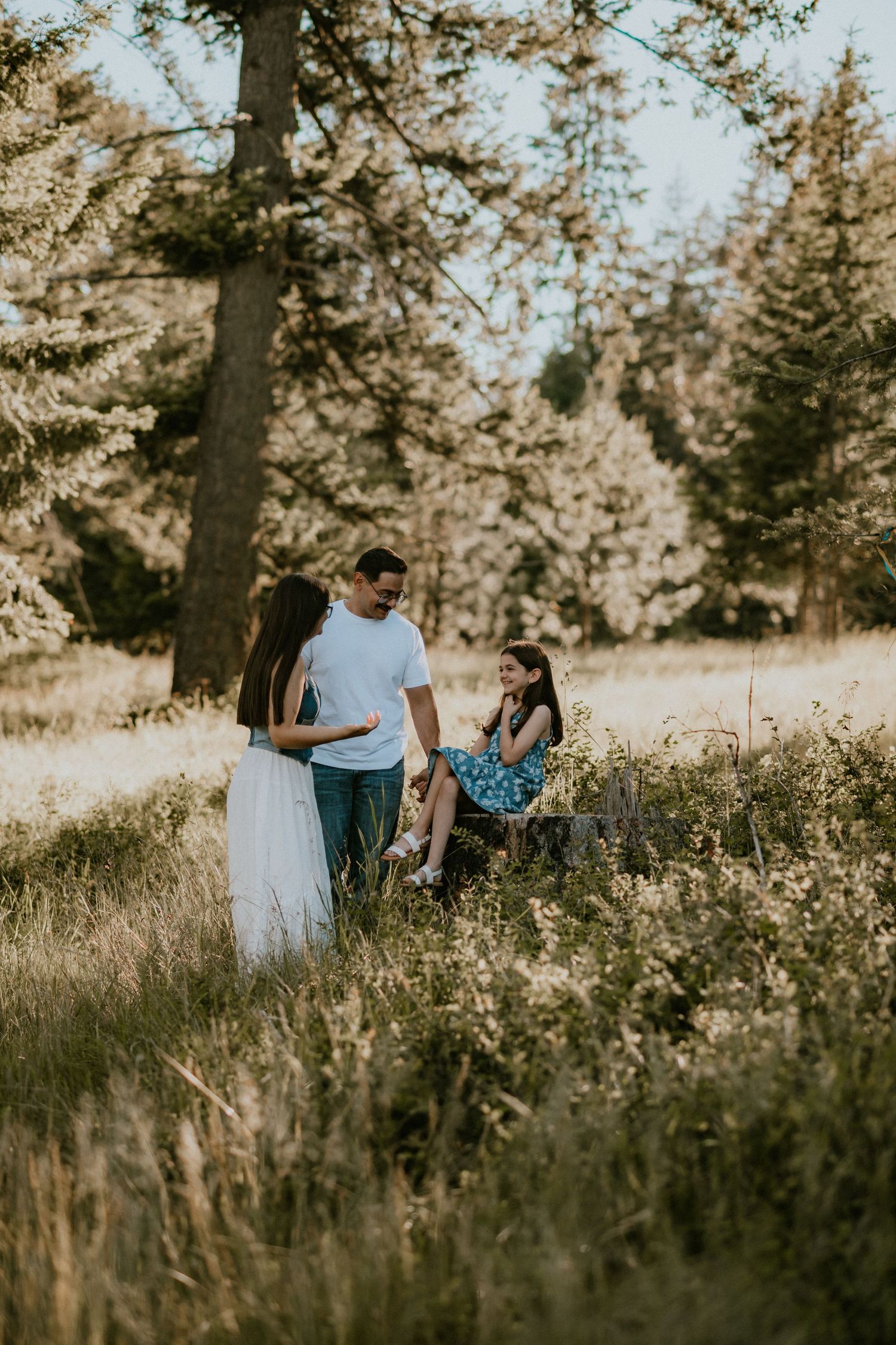 Family of three enjoying a sunny outdoor moment in a meadow surrounded by tall pine trees and wildflowers.