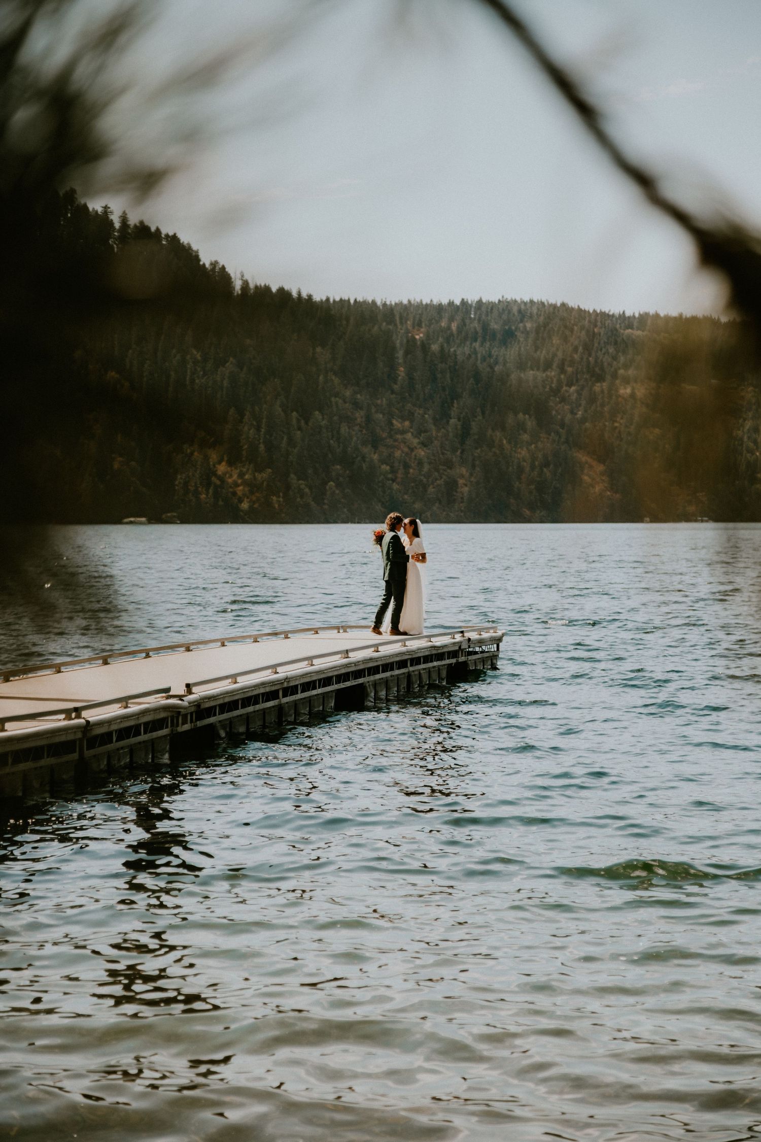 A couple stands together on a wooden dock overlooking a serene mountain lake surrounded by evergreen forest.