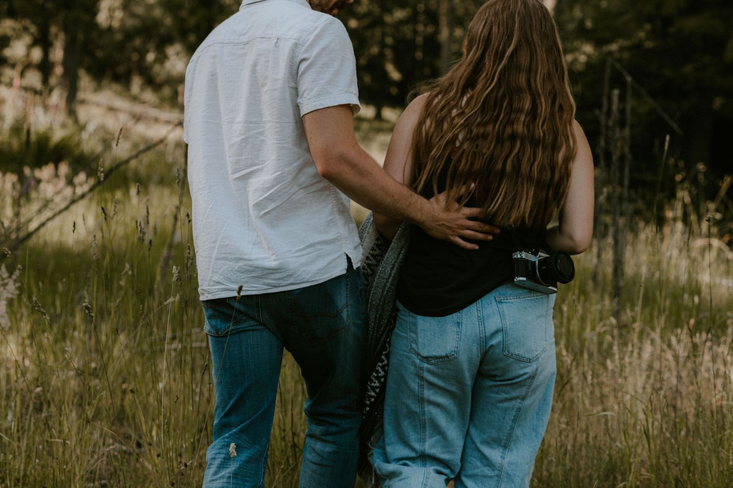 A couple in jeans and white shirts embracing in a sunlit field of tall grass and wildflowers.