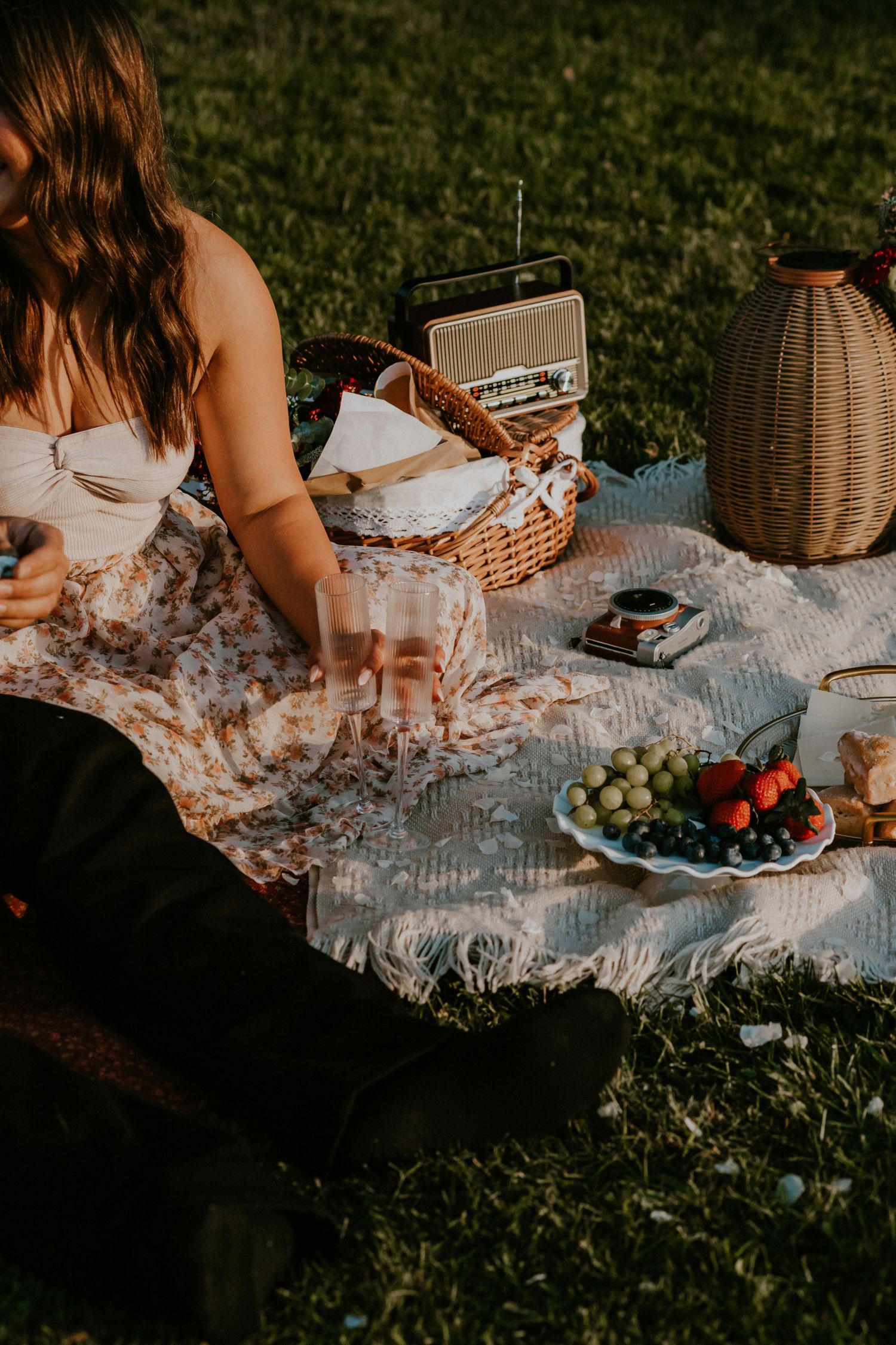 A picnic setup on a white blanket with various foods, fruits, snacks, and a wicker basket laid out on green grass.