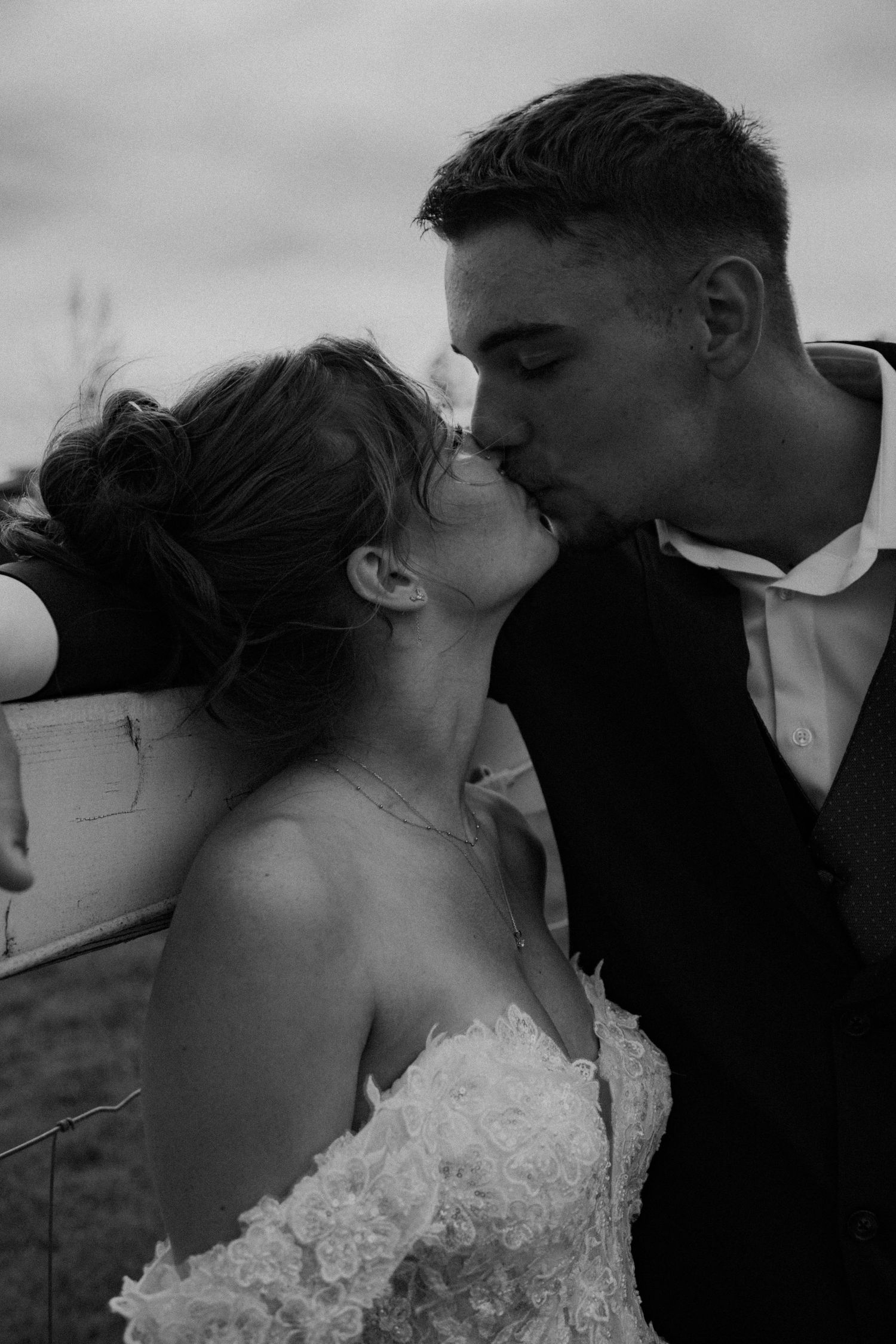 Black and white romantic wedding portrait showing couple kissing outdoors, with bride in lace dress and groom in formal suit.