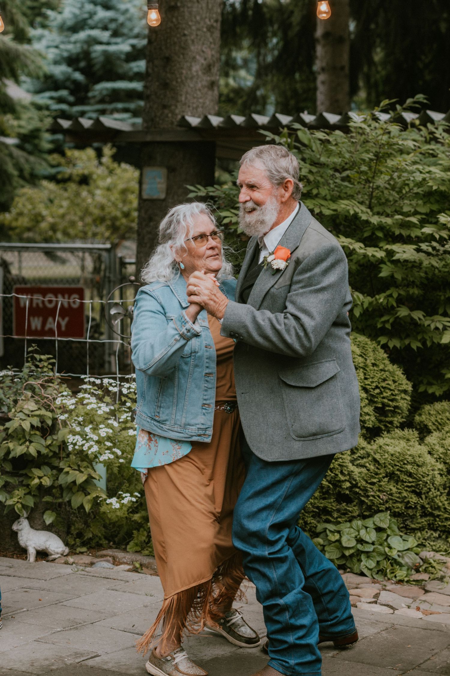 Couple dancing together outdoors in garden setting with string lights and evergreen trees in background.