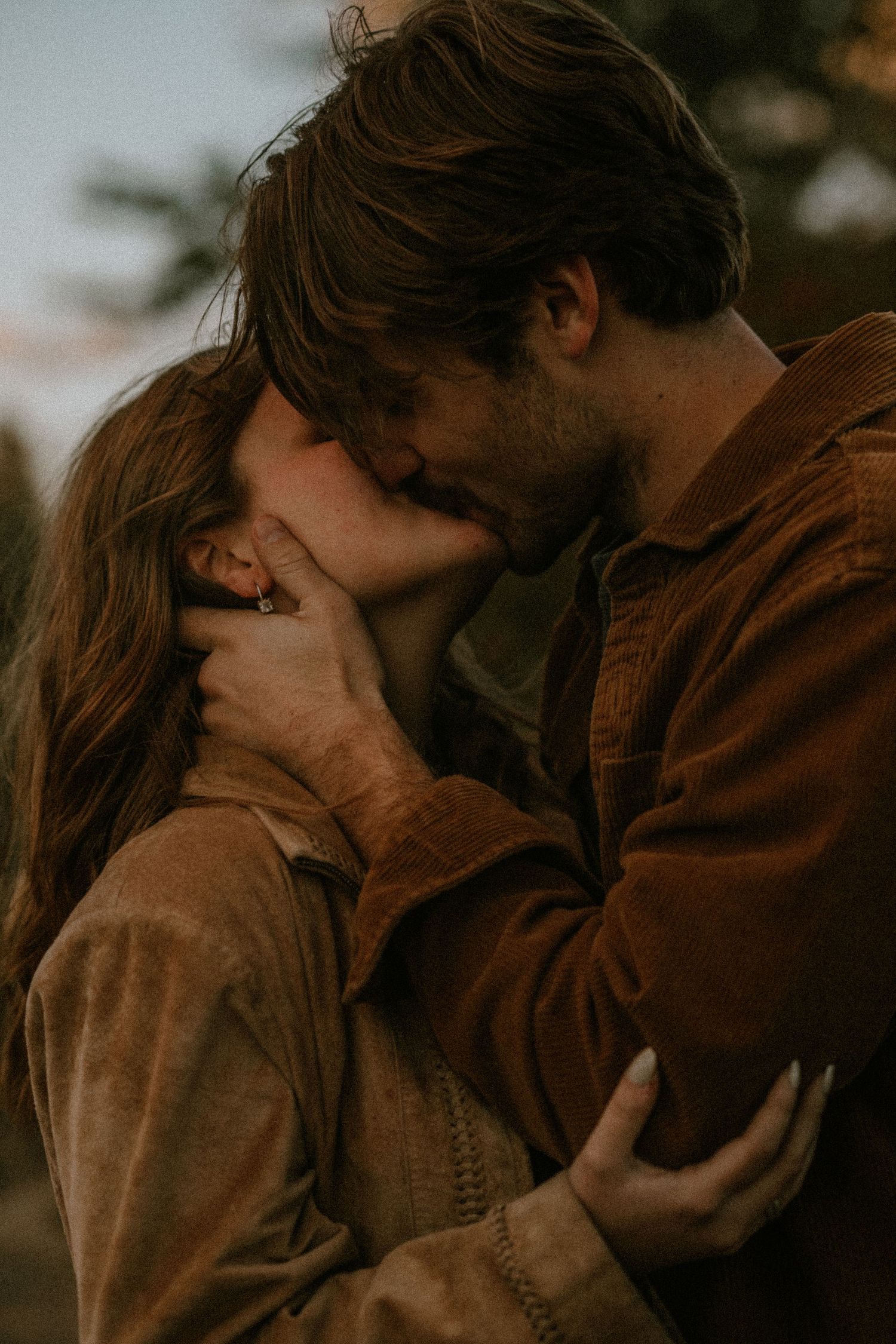 A couple sharing a romantic kiss outdoors, both wearing matching brown corduroy jackets in warm golden light.
