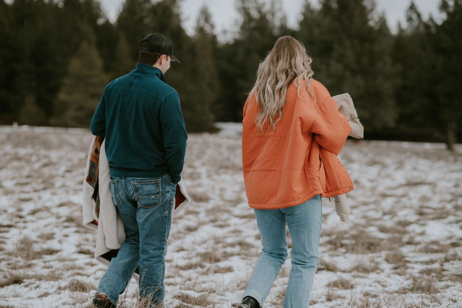 A couple walks hand in hand through a snowy winter landscape surrounded by evergreen trees in the background.