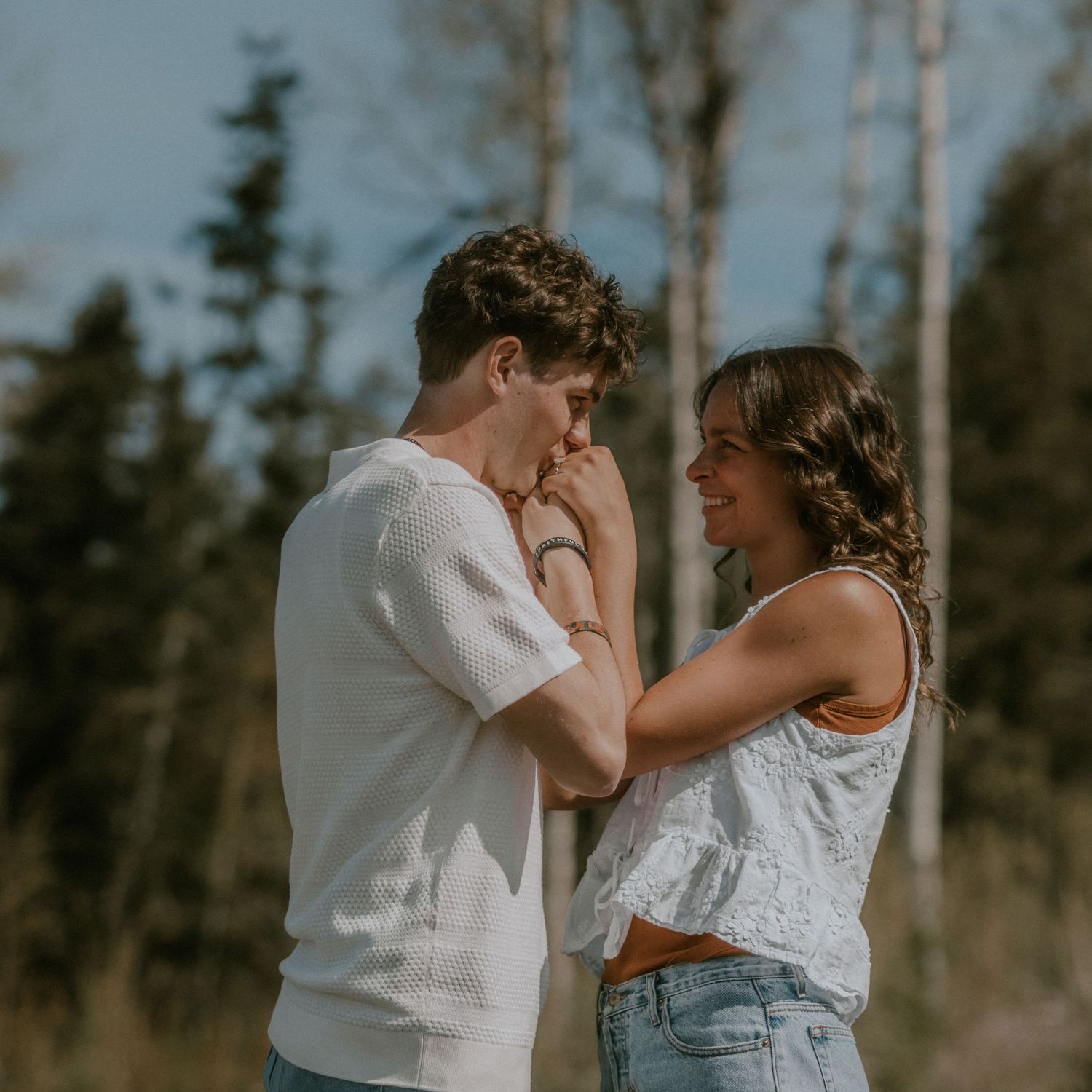 A young couple shares a romantic moment outdoors, the man kissing her hand while she smiles, with trees in the background.
