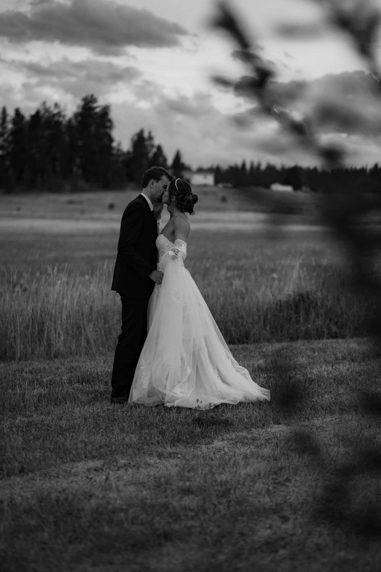 Black and white wedding photo of a couple walking together through a grassy meadow captured through branches and leaves.