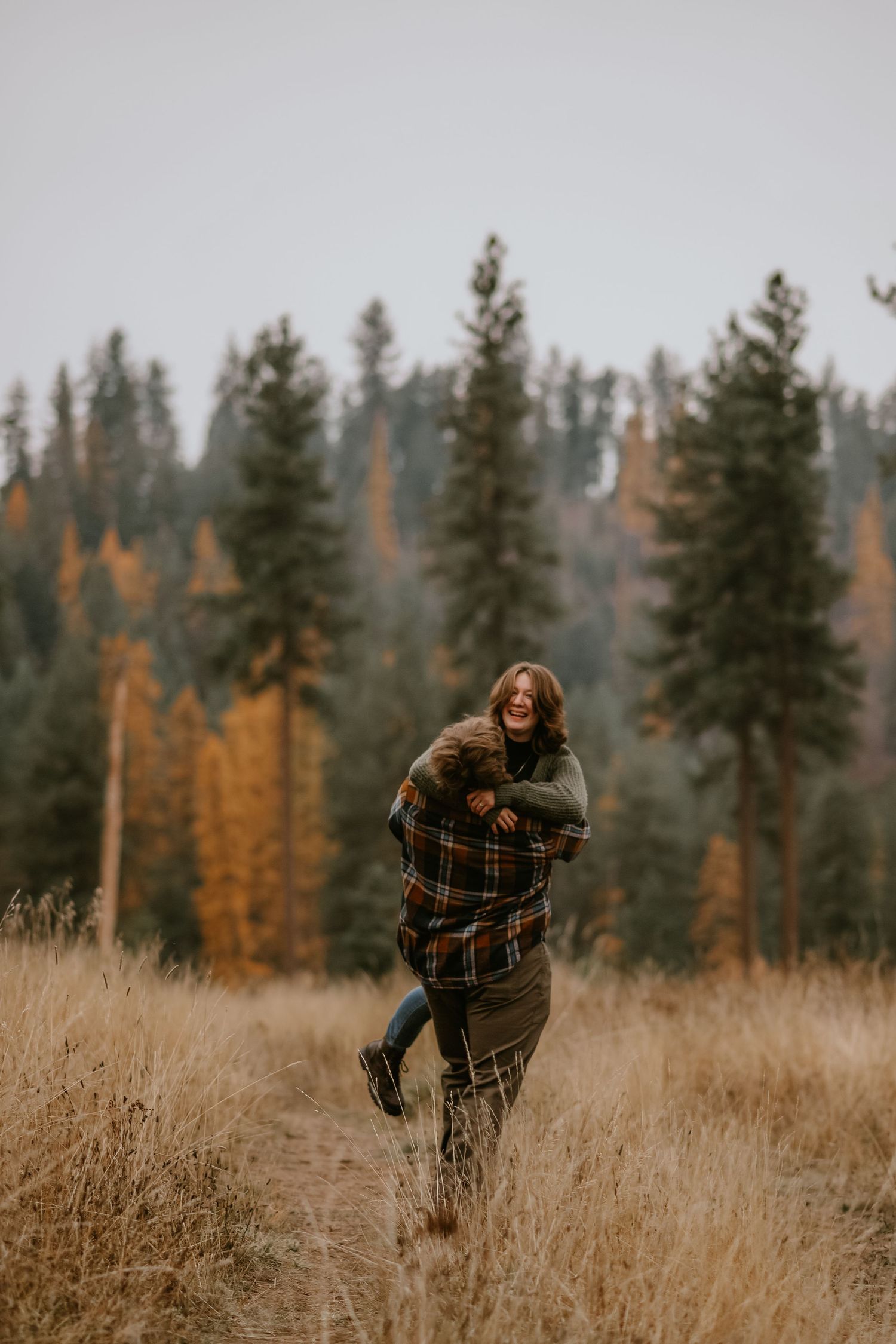 A person wearing a plaid shirt dances and jumps through a golden wheat field with autumn evergreen trees in the distance.