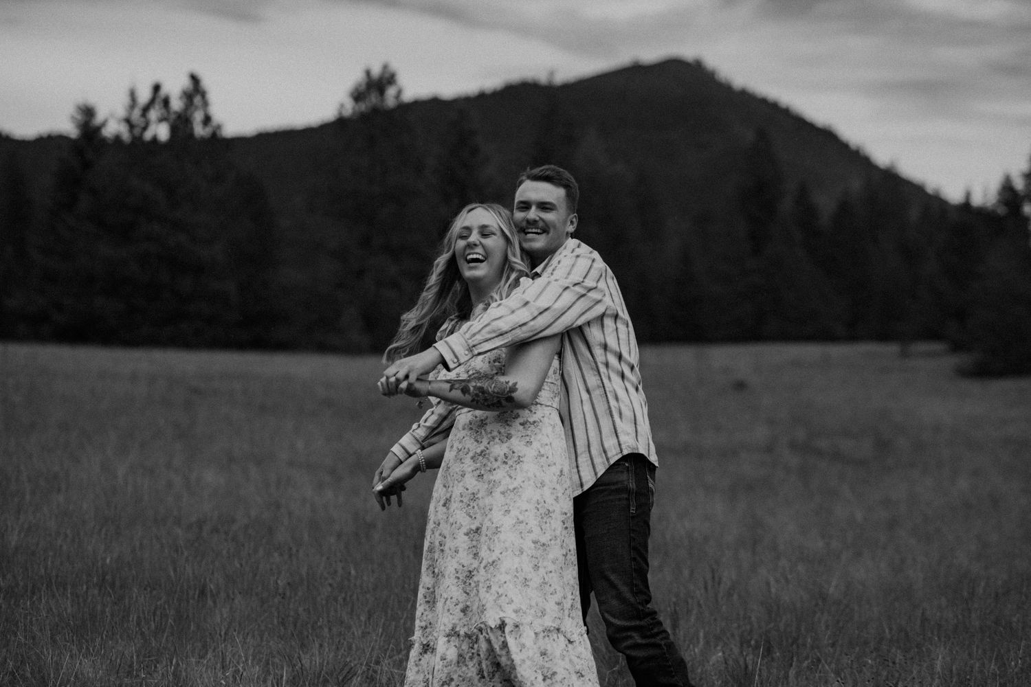 A couple embraces in a meadow against a mountainous backdrop in black and white photography.