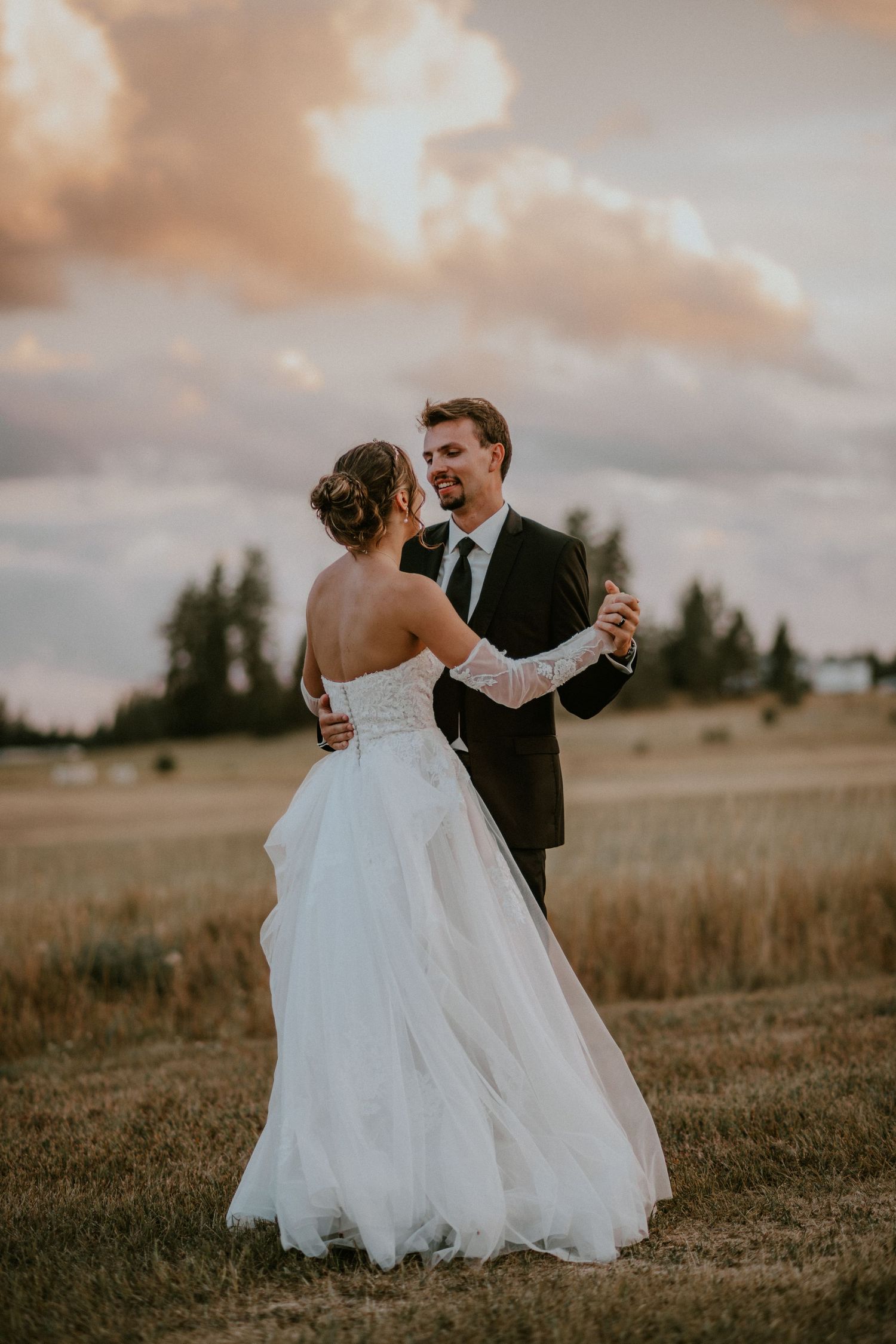 Newlywed couple dances romantically in a golden field during sunset while bride's white dress flows in the wind.