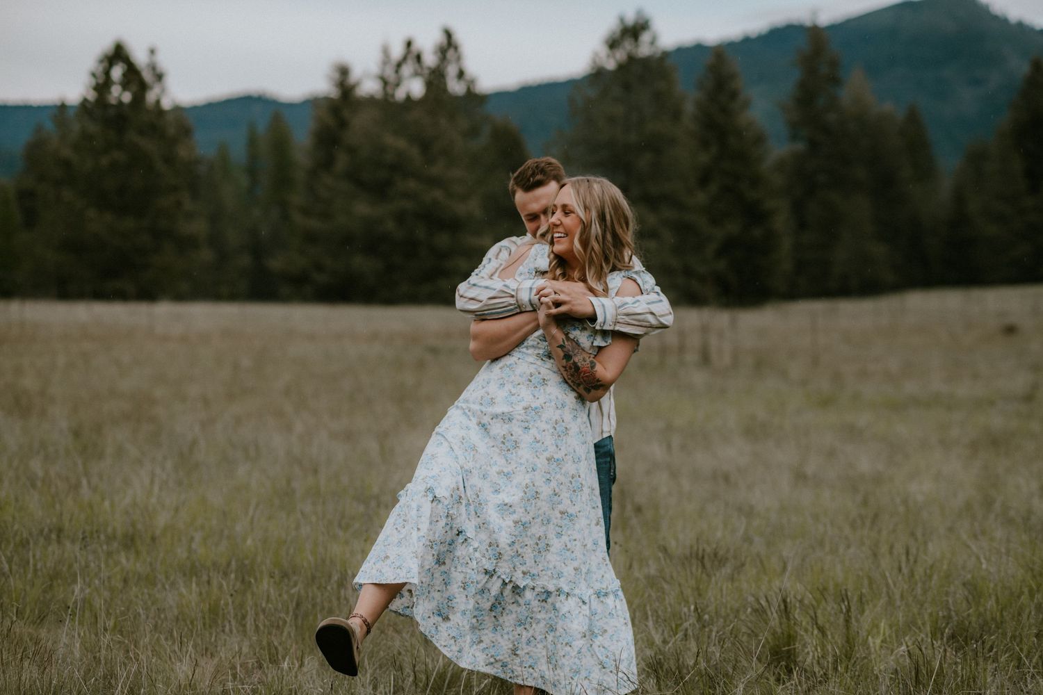 Person in floral dress spins joyfully in a mountain meadow with evergreen trees and mountains in the background.