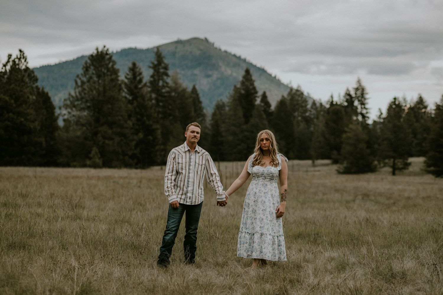 A romantic couple holds hands in a scenic mountain meadow during their engagement photo session.