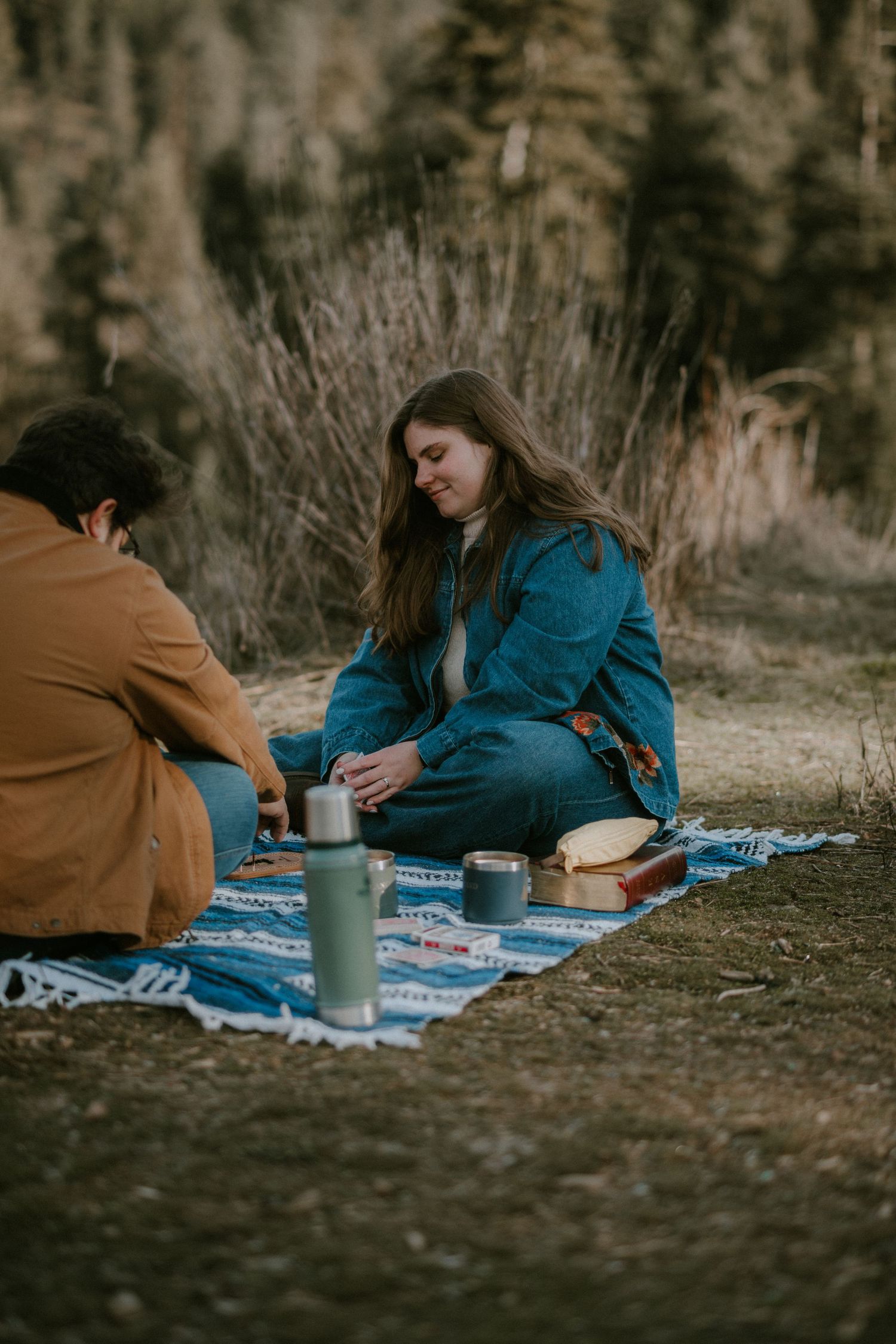 Two people enjoy an outdoor picnic on a blue blanket with Stanley thermoses, surrounded by nature in cool weather.