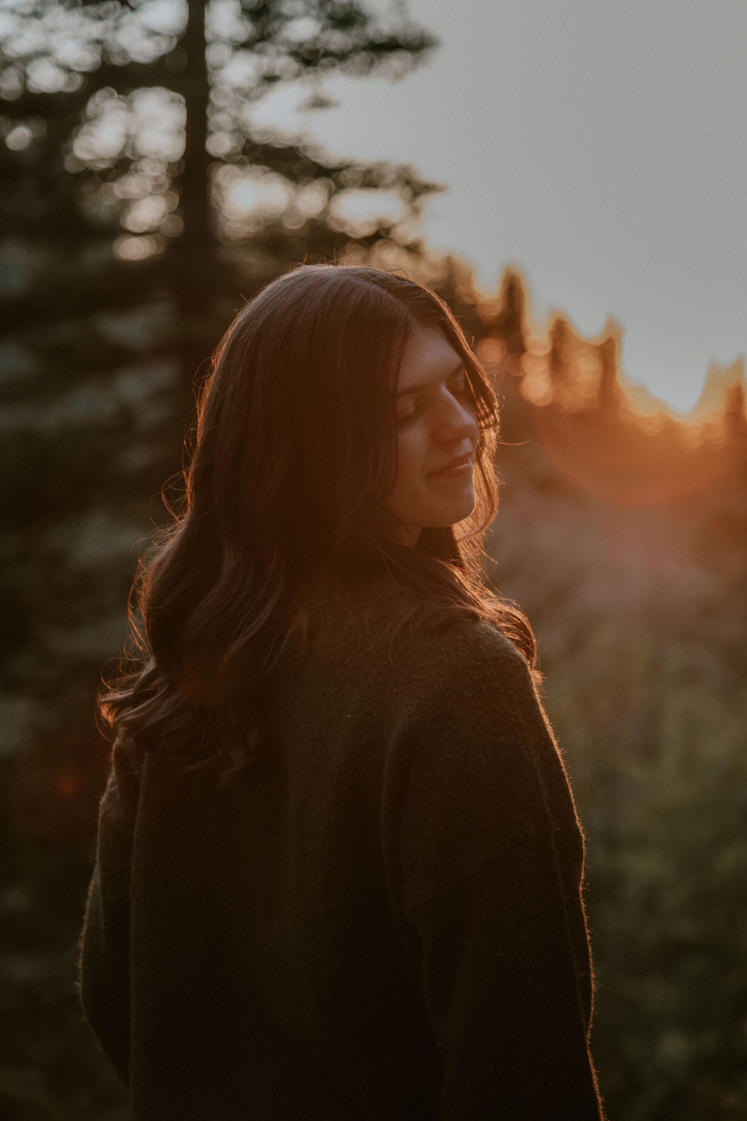 Silhouette of a person with long flowing hair backlit by a warm sunset through trees creating a golden rim light effect.
