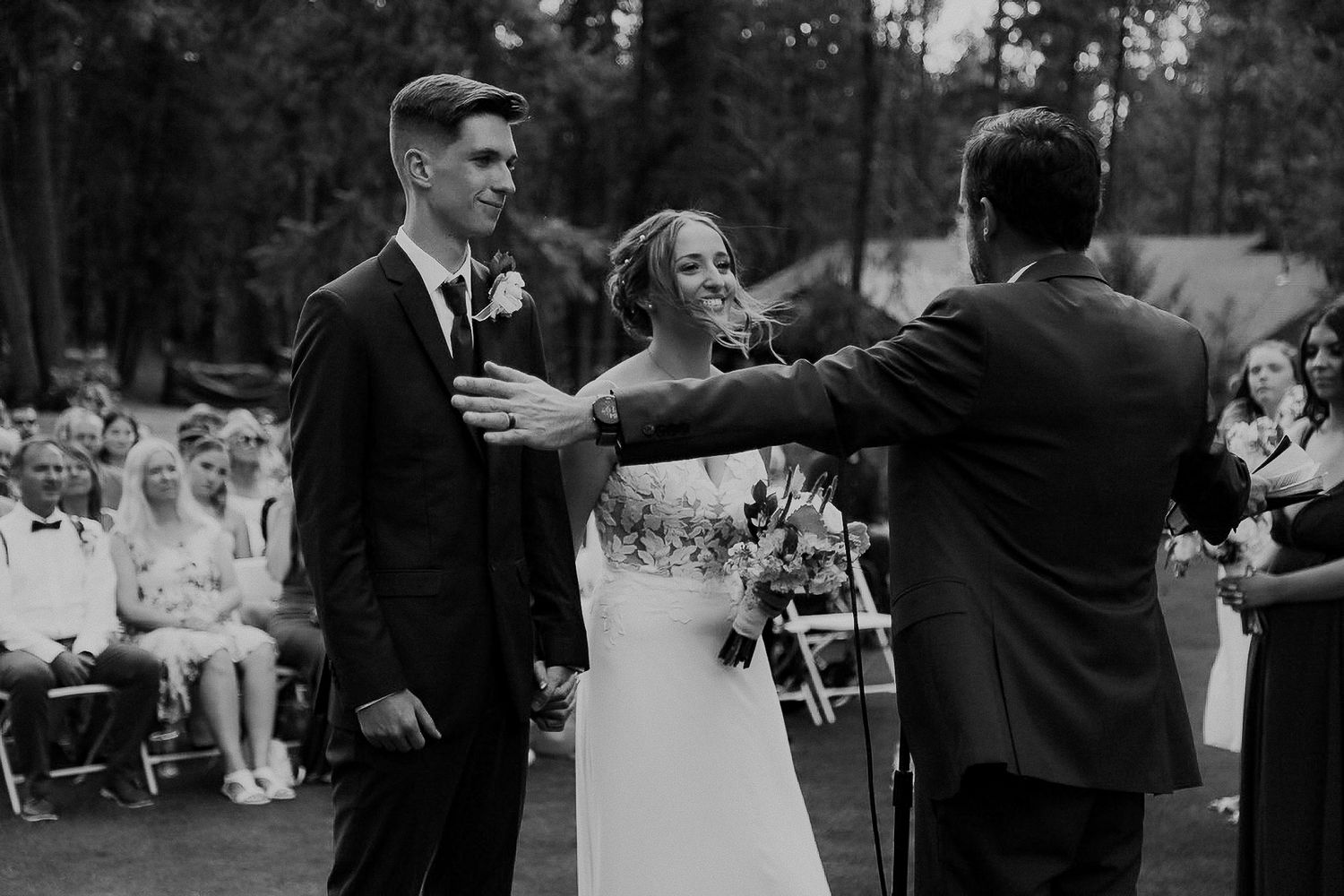 Black and white photo of a joyful outdoor wedding ceremony moment with people celebrating in a wooded setting.