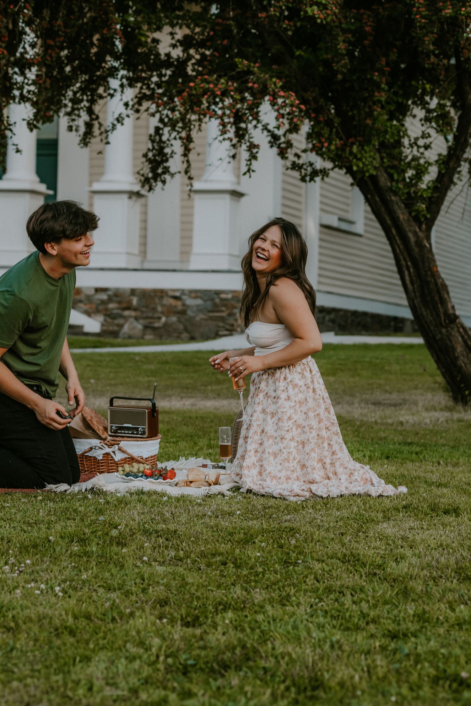 A couple enjoys a romantic summer picnic on the lawn with a vintage radio and snacks under a tree.