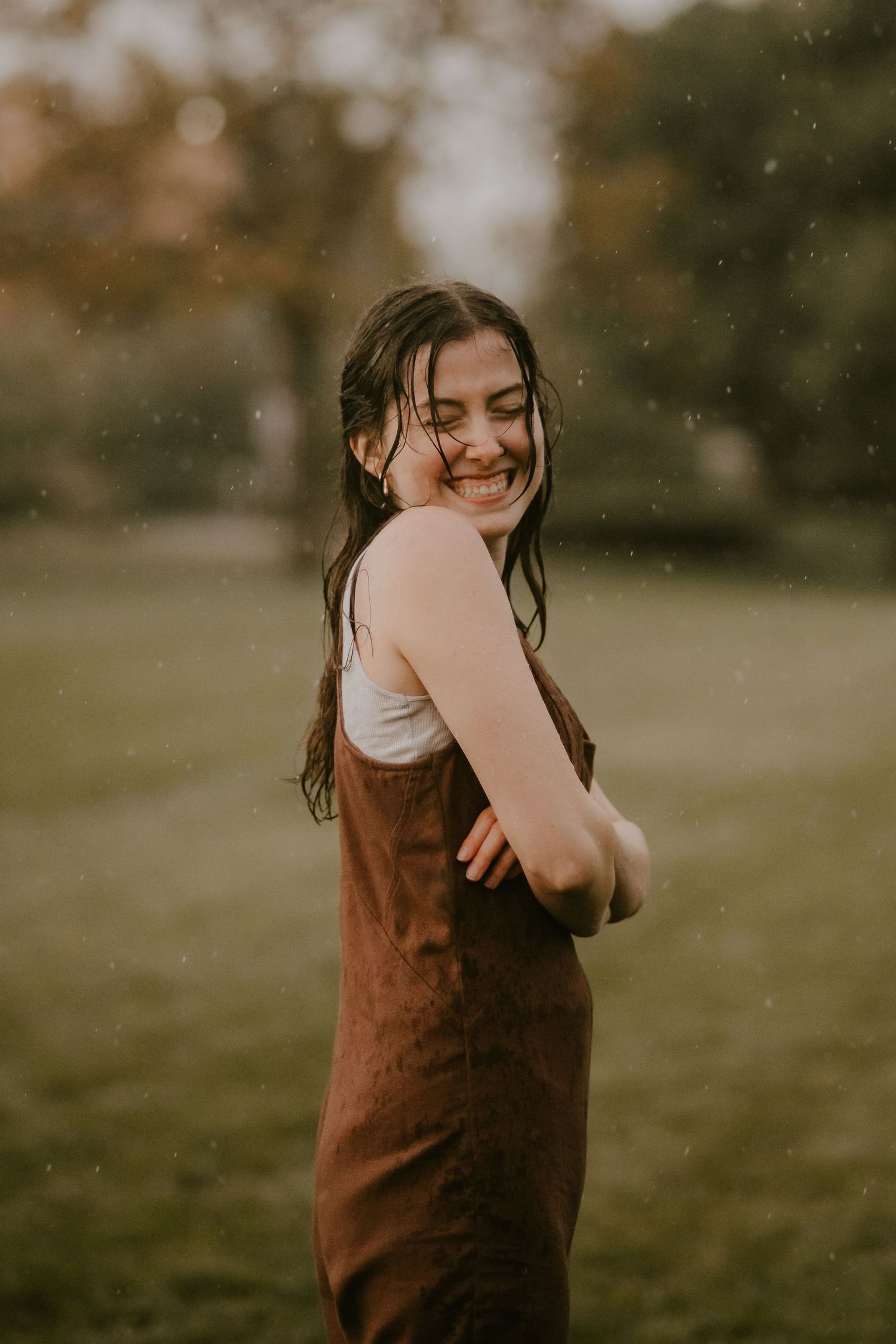 A woman in a brown dress laughs joyfully while standing in a grassy field on a warm autumn day.