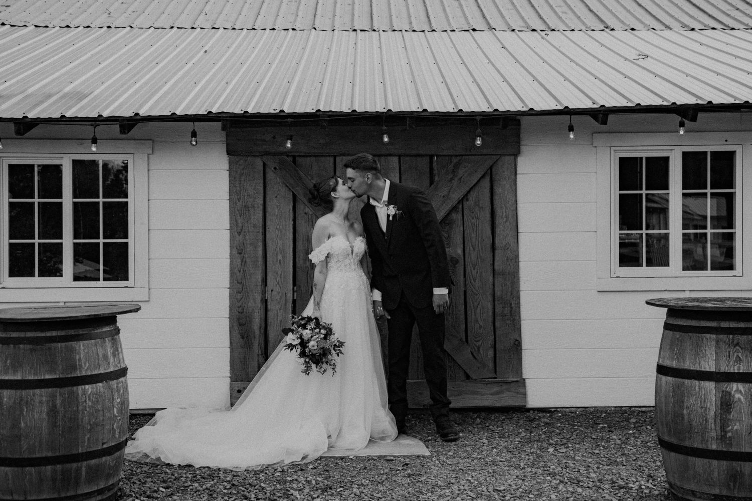 A romantic black and white photograph of a couple sharing an intimate moment outside a rustic barn with wine barrels.